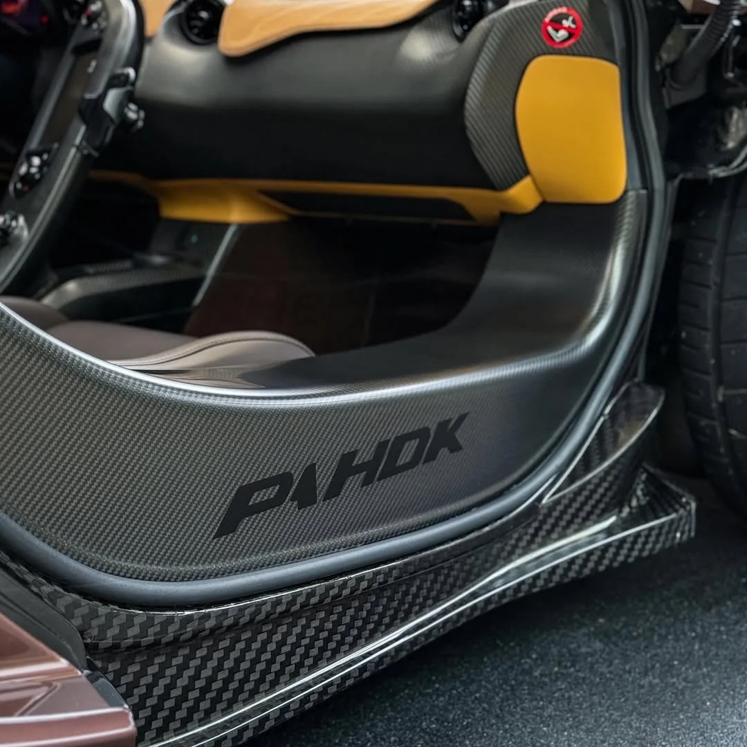 Close-up of a carbon fiber side panel of a car with the word 'PAHOK' on it, showing a yellow and black interior with a small sticker on the dashboard, and part of the tire visible.