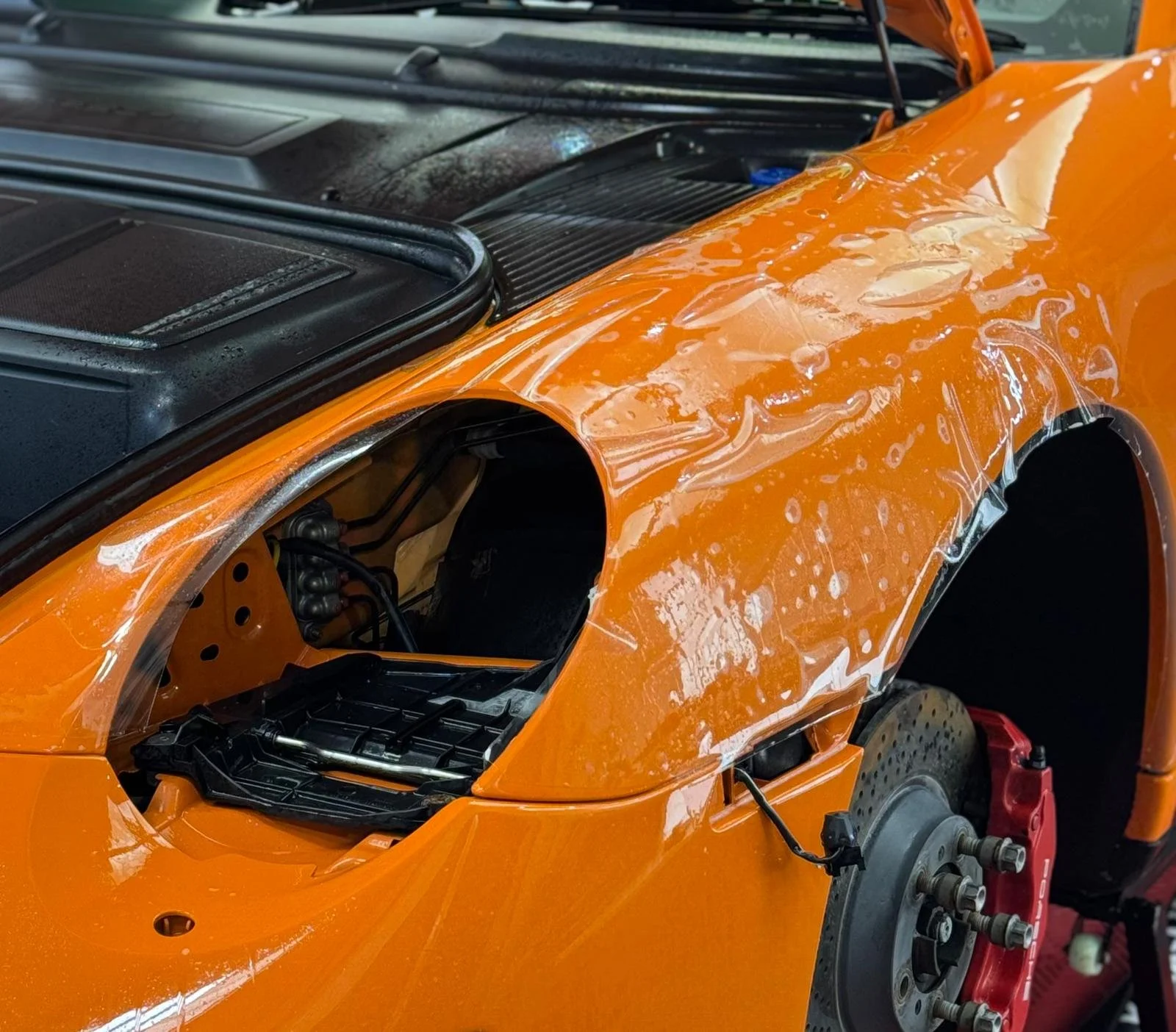 Close-up of an orange sports car in the process of being repaired or assembled, with the front wheel removed and the wheel well exposed.