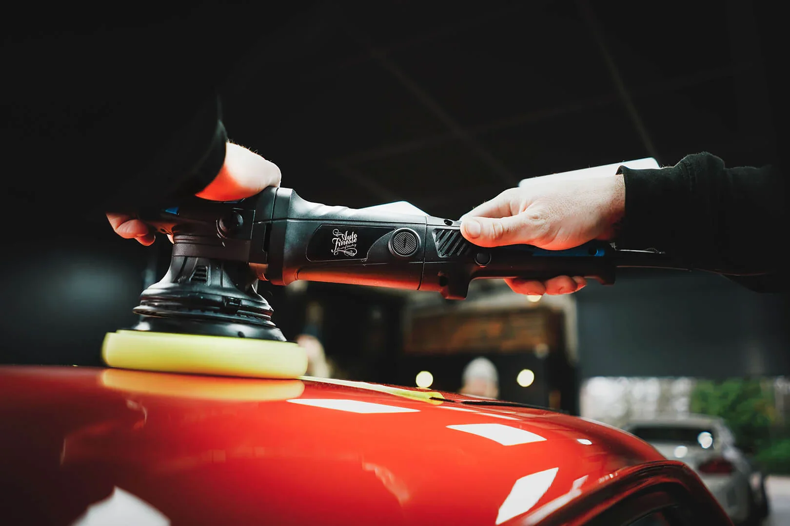 Person polishing a red car with a polishing machine in an indoor garage.