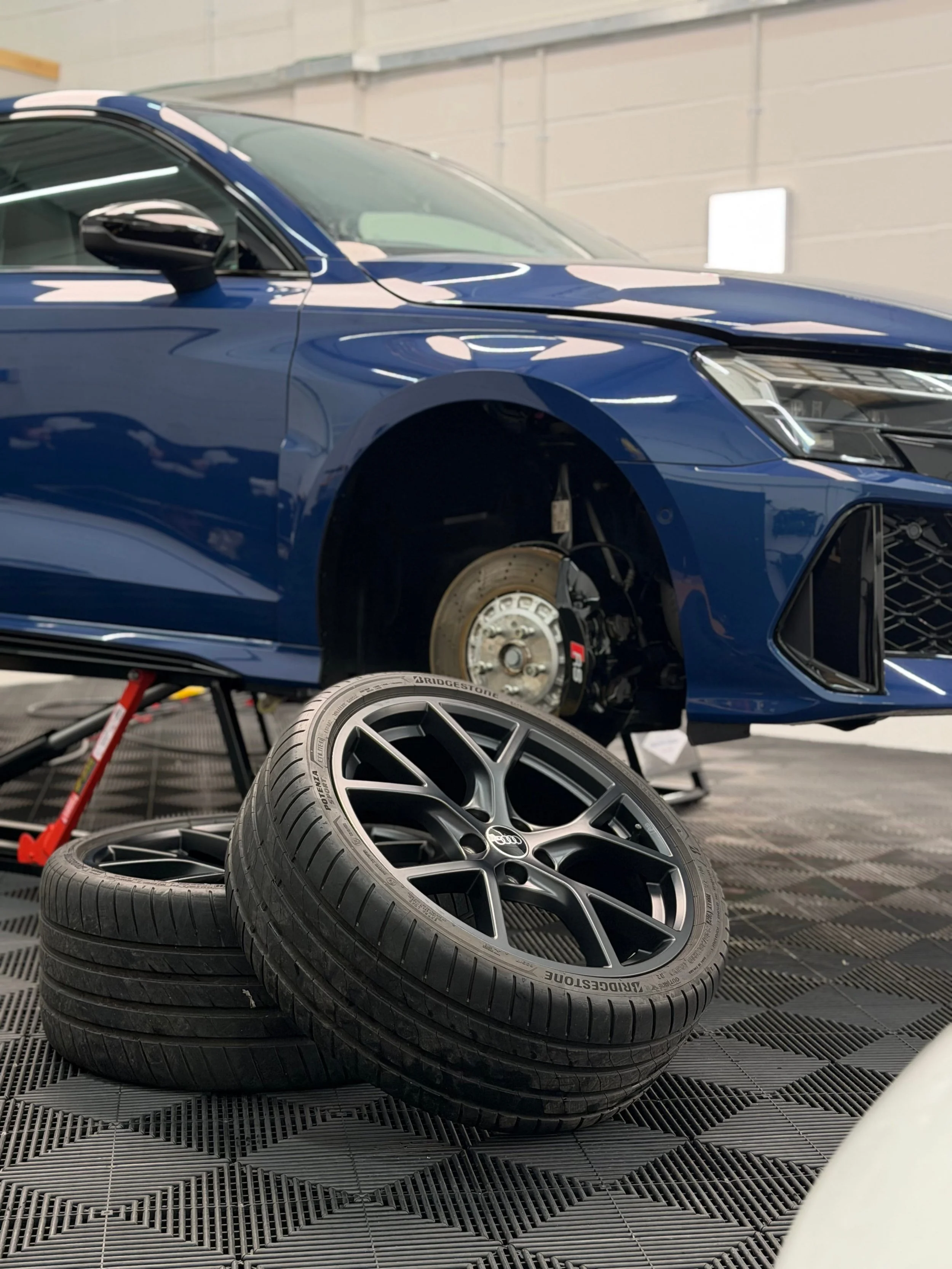 Regenerate alt text copyIconcrossIconBlue A blue car is lifted for maintenance with its front wheel removed; vehicle detailing supplies and alloy wheels are stacked nearby.