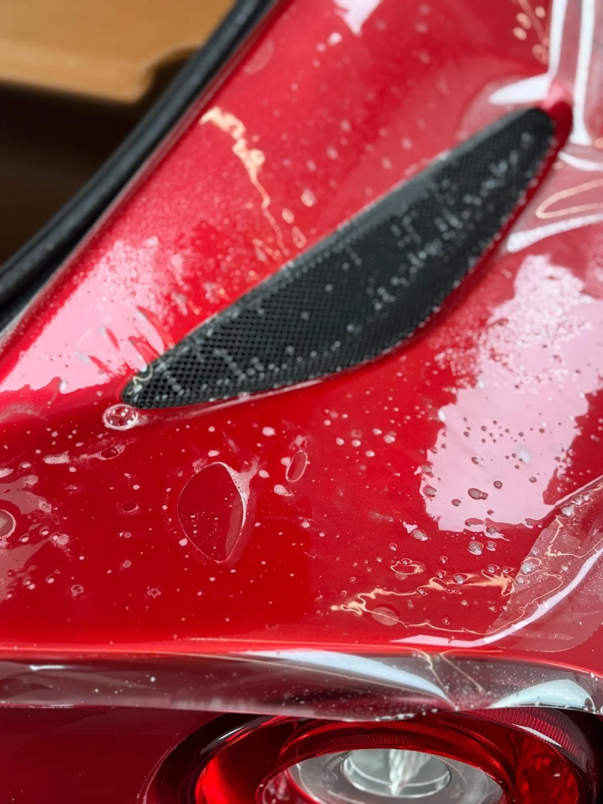 Close-up of a red car roof with a black roof vent, water droplets, and soap suds on the surface.