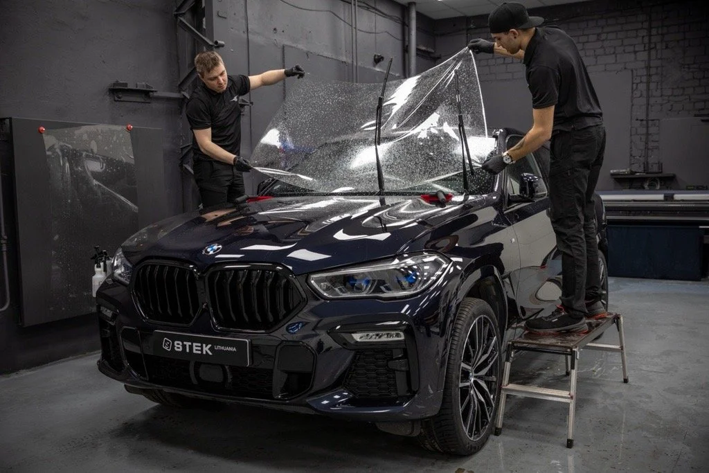 Two men applying Stek Dyno Top protective film to the windshield of a black BMW SUV inside an auto workshop.