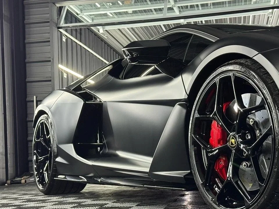 A black and silver luxury sports car parked in a garage, showing a close-up of the front left side, including the wheel with a red brake caliper.