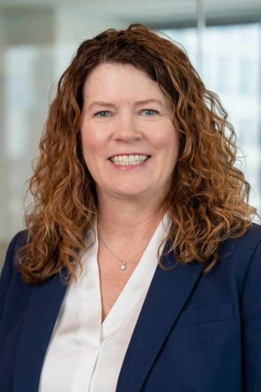 A woman with curly red hair dressed in a navy blazer and white blouse, smiling in a professional office setting.