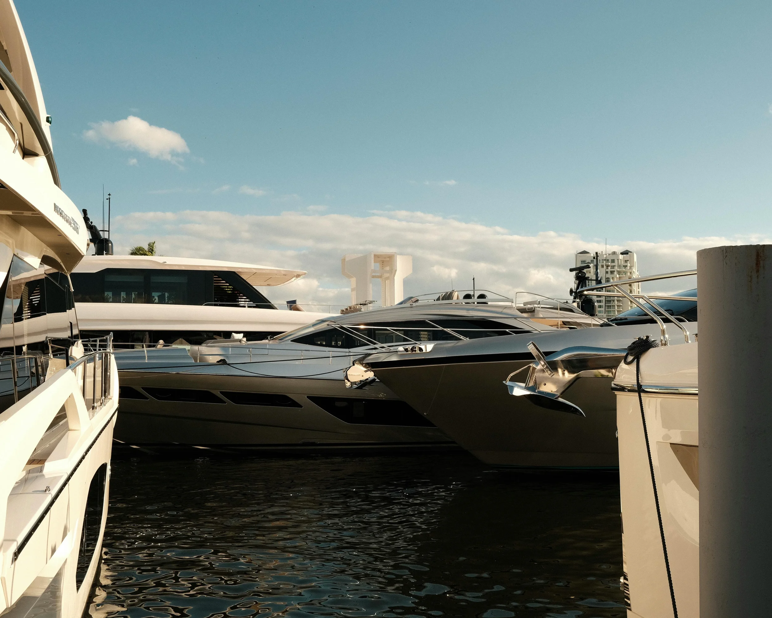 A marina with several yachts and boats docked, with high-rise buildings in the background and a partly cloudy sky.