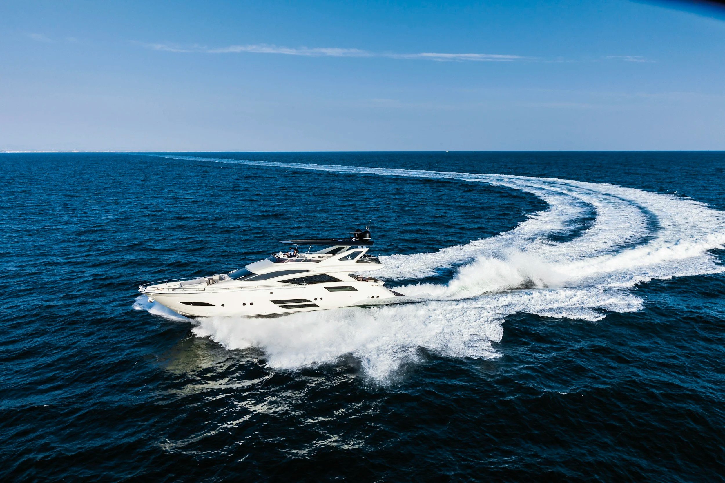 A white yacht moving swiftly across the blue ocean, creating a wake behind it with a clear blue sky overhead.