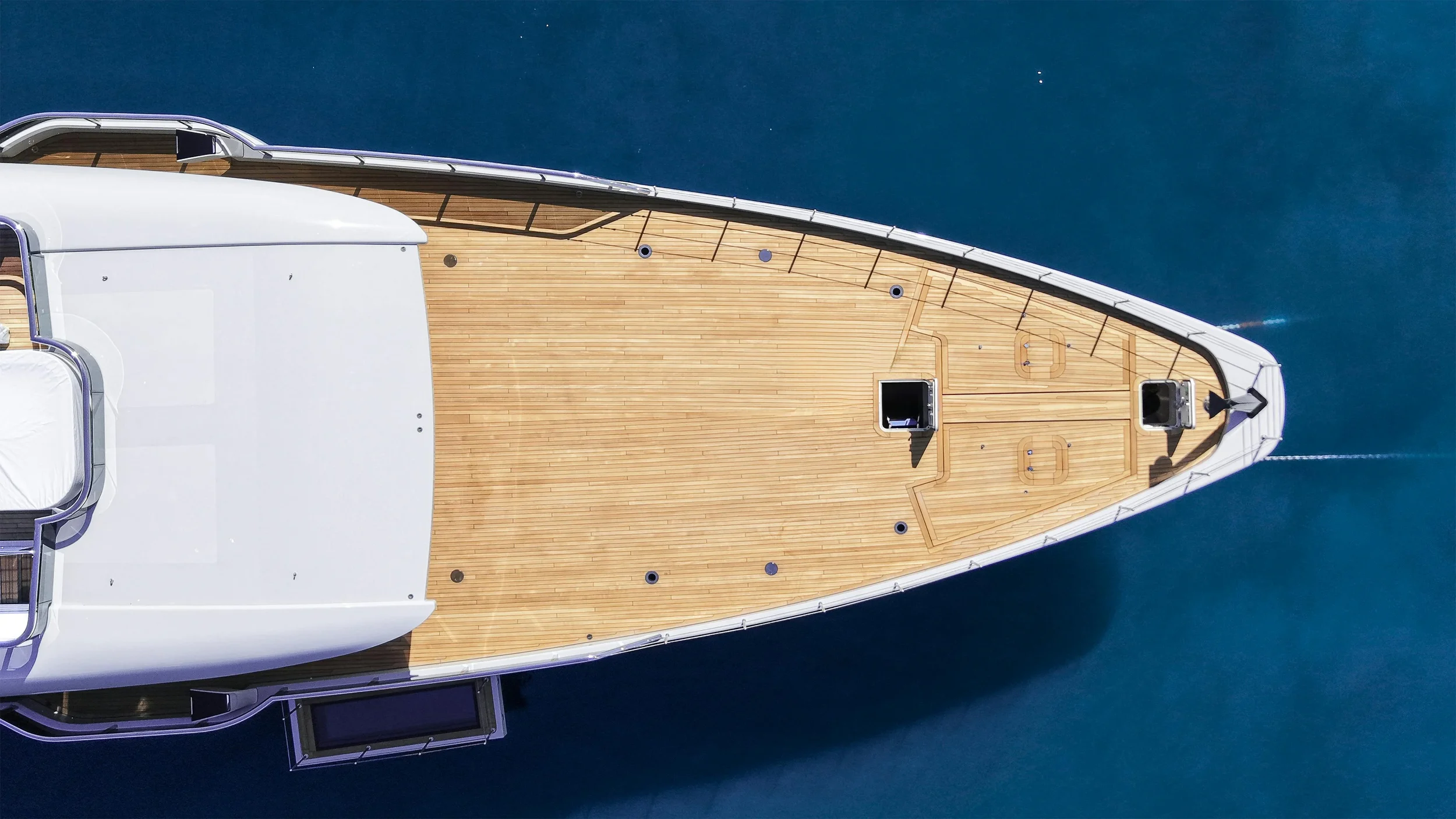 Top-down aerial view of the bow of a yacht with a wooden deck, two hatches, and the surrounding blue water.