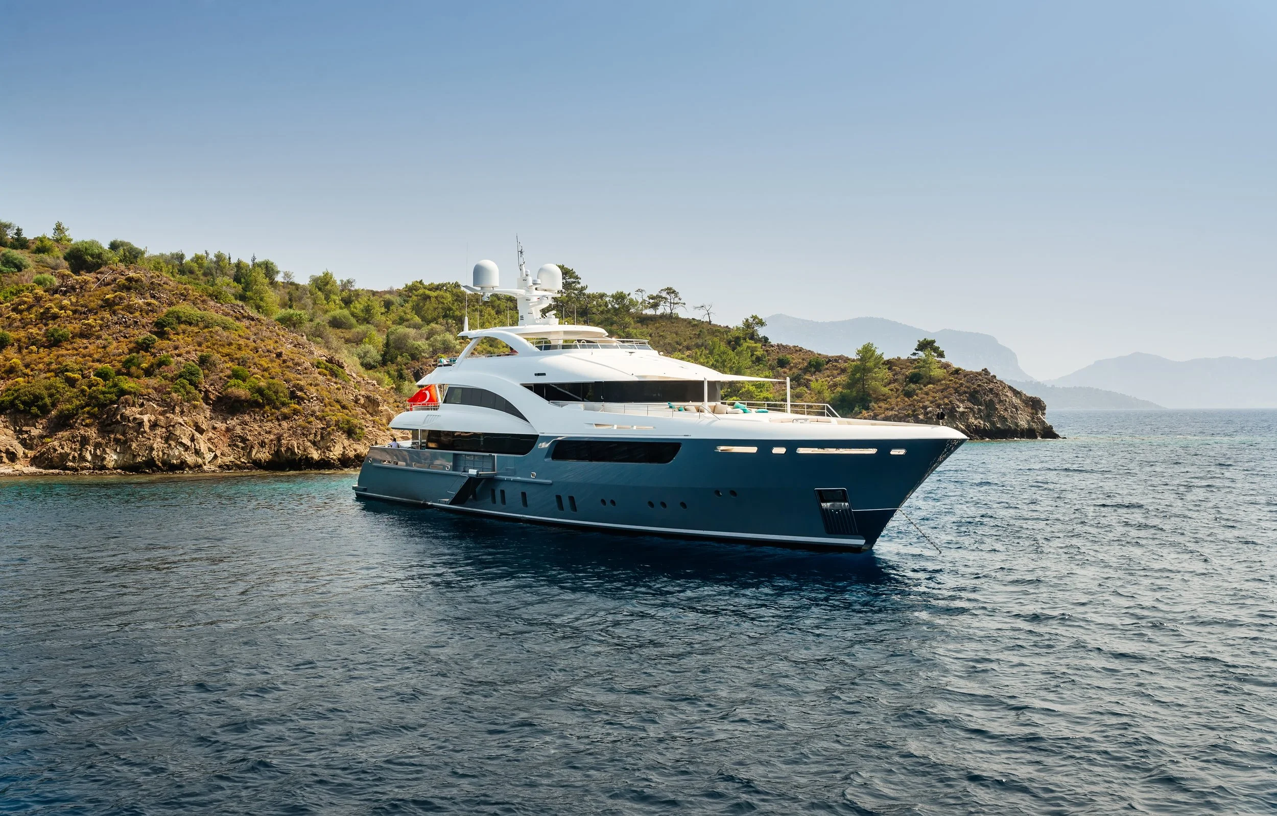 A white yacht in water near a cliff with green trees, with distant mountains under a clear blue sky.