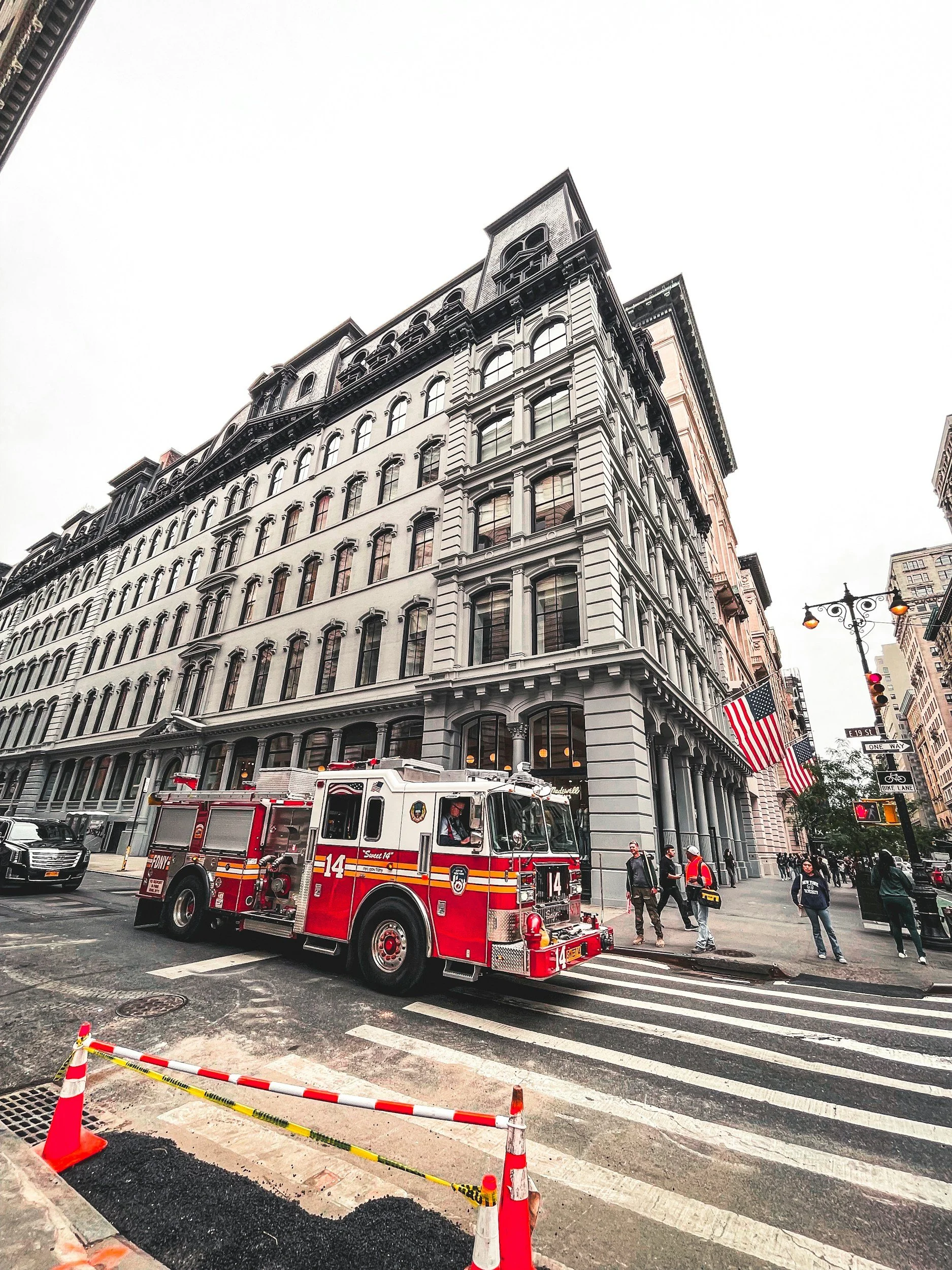 A red fire truck driving down a city street in front of a large historic building with arch windows and a detailed facade. There are pedestrians on the sidewalk and traffic signals visible in the scene.