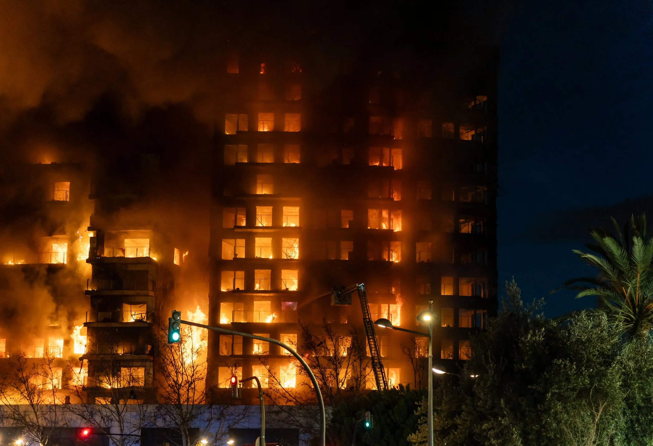 A multi-story building is engulfed in flames and thick smoke during a fire at night. Firefighters are actively battling the blaze, with some equipment visible at the scene. The background is dark, and a traffic light is lit green in the foreground.