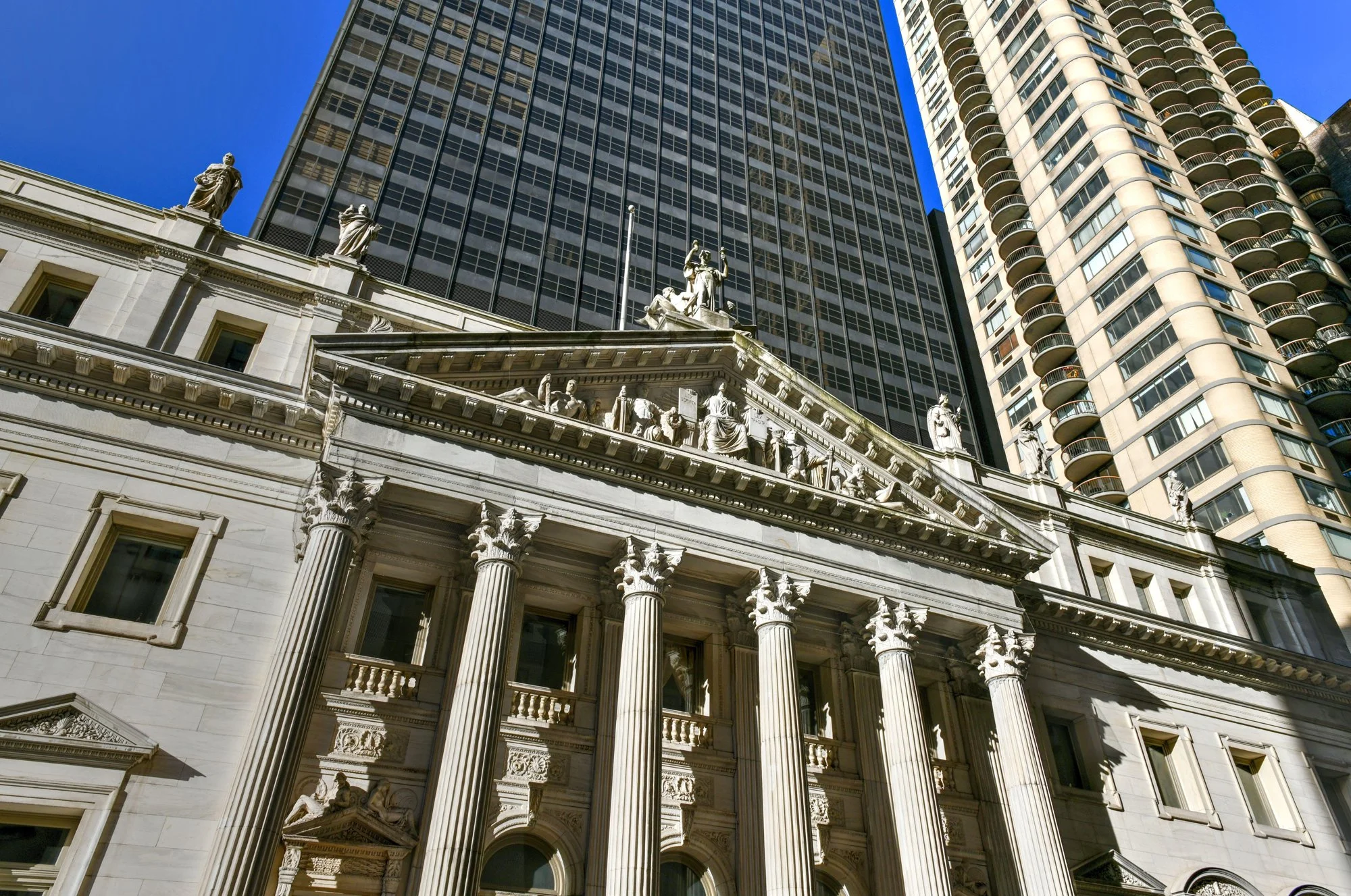 A close-up view of a historic building with ornate classical architecture featuring tall columns and sculptures on the facade, with modern high-rise buildings in the background and a clear blue sky.