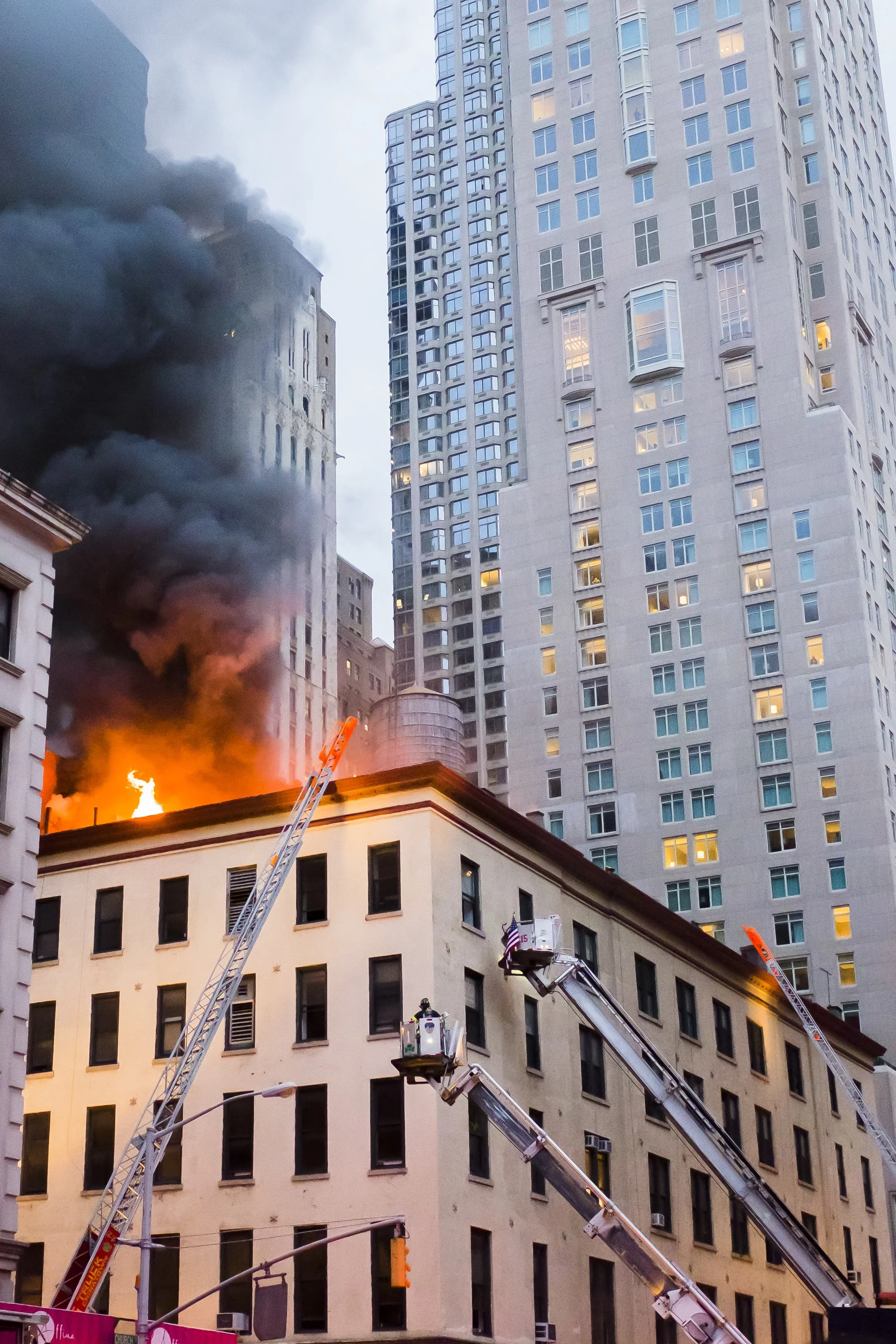 A building on fire with black smoke billowing out of the windows. Firefighters are using ladders and a fire truck to combat the flames in an urban area with tall skyscrapers.