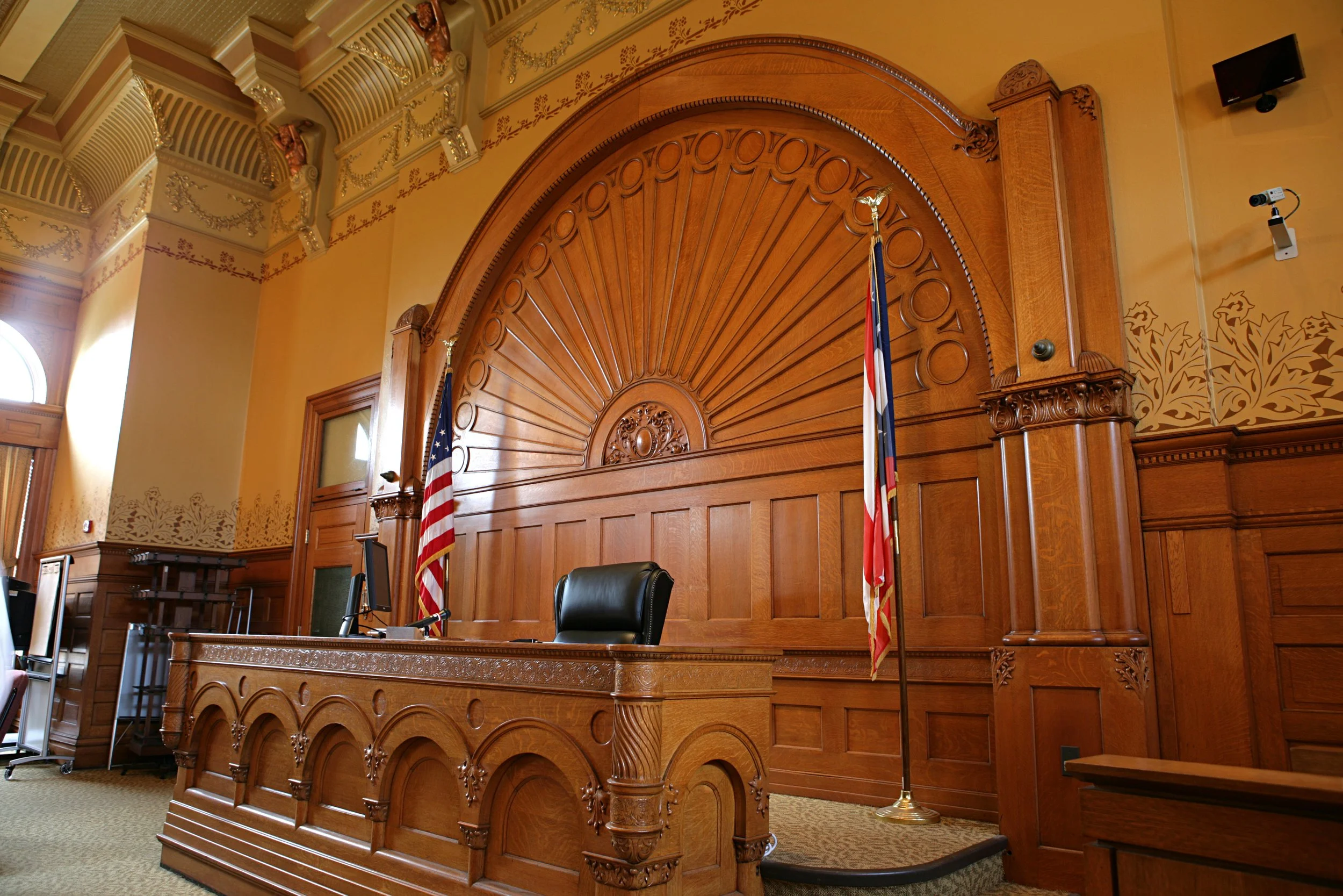A courtroom with a large, ornate wooden wall behind a judge's bench. Flags are positioned on either side of the bench. There are security cameras and a monitor visible.