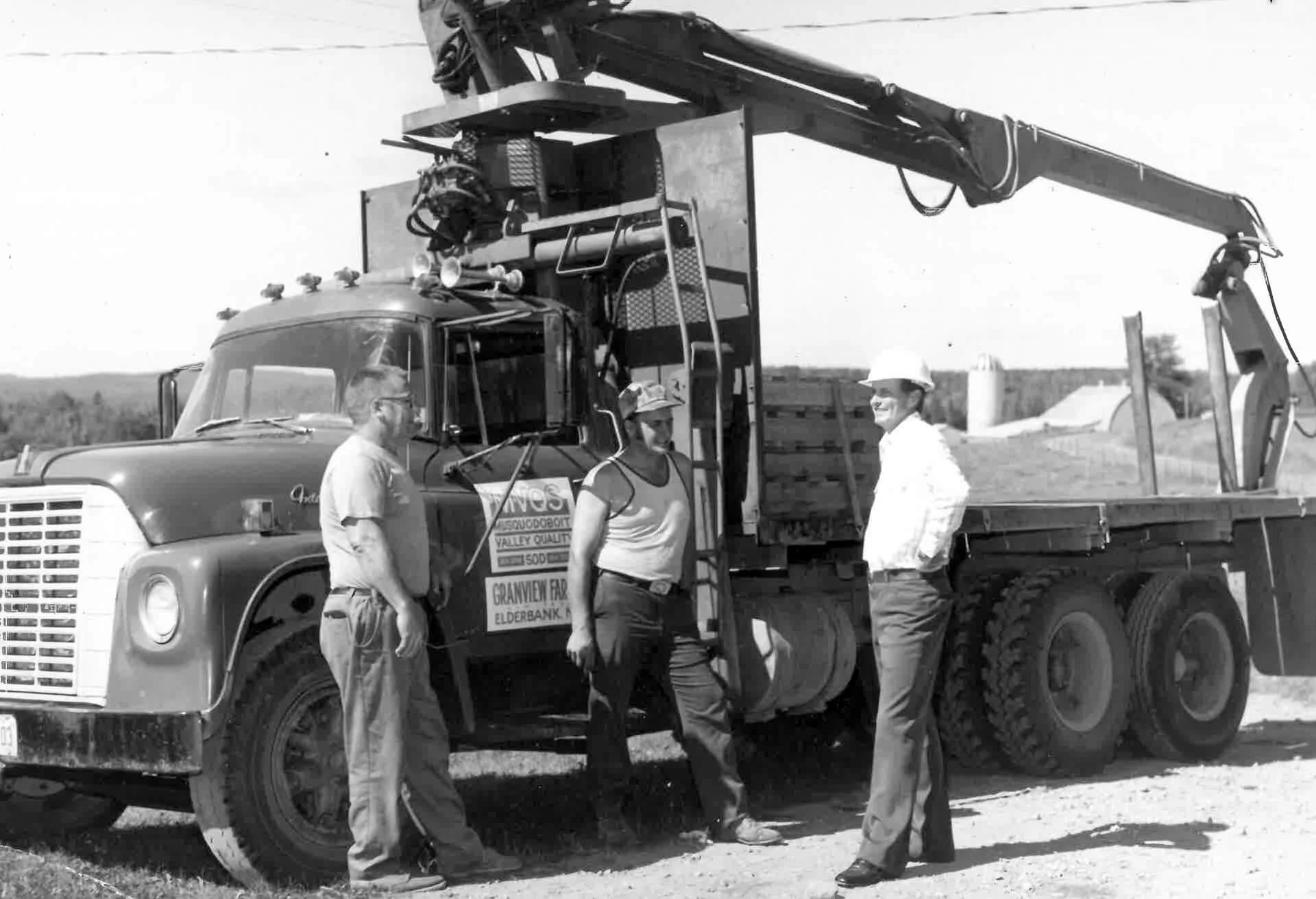Three men stand beside a large truck with a mounted crane. The men are engaged in conversation, with two wearing hats and one wearing sunglasses. The truck has a sign on the door, and the background appears to be a rural or construction area with som