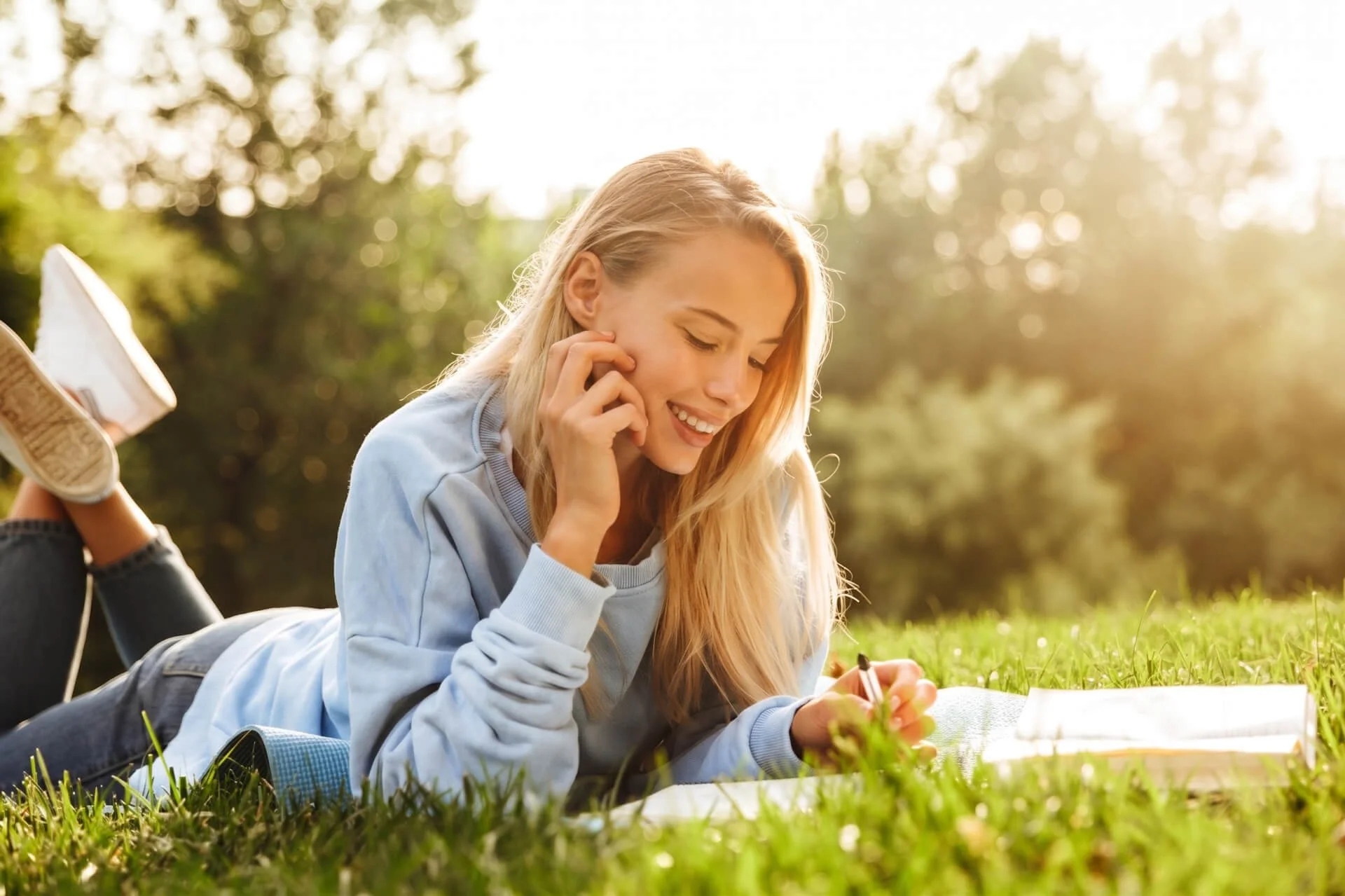 A young woman with long blonde hair lying on her stomach on the grass, smiling, with a notebook and pen nearby, outdoors in sunlight with trees in the background.