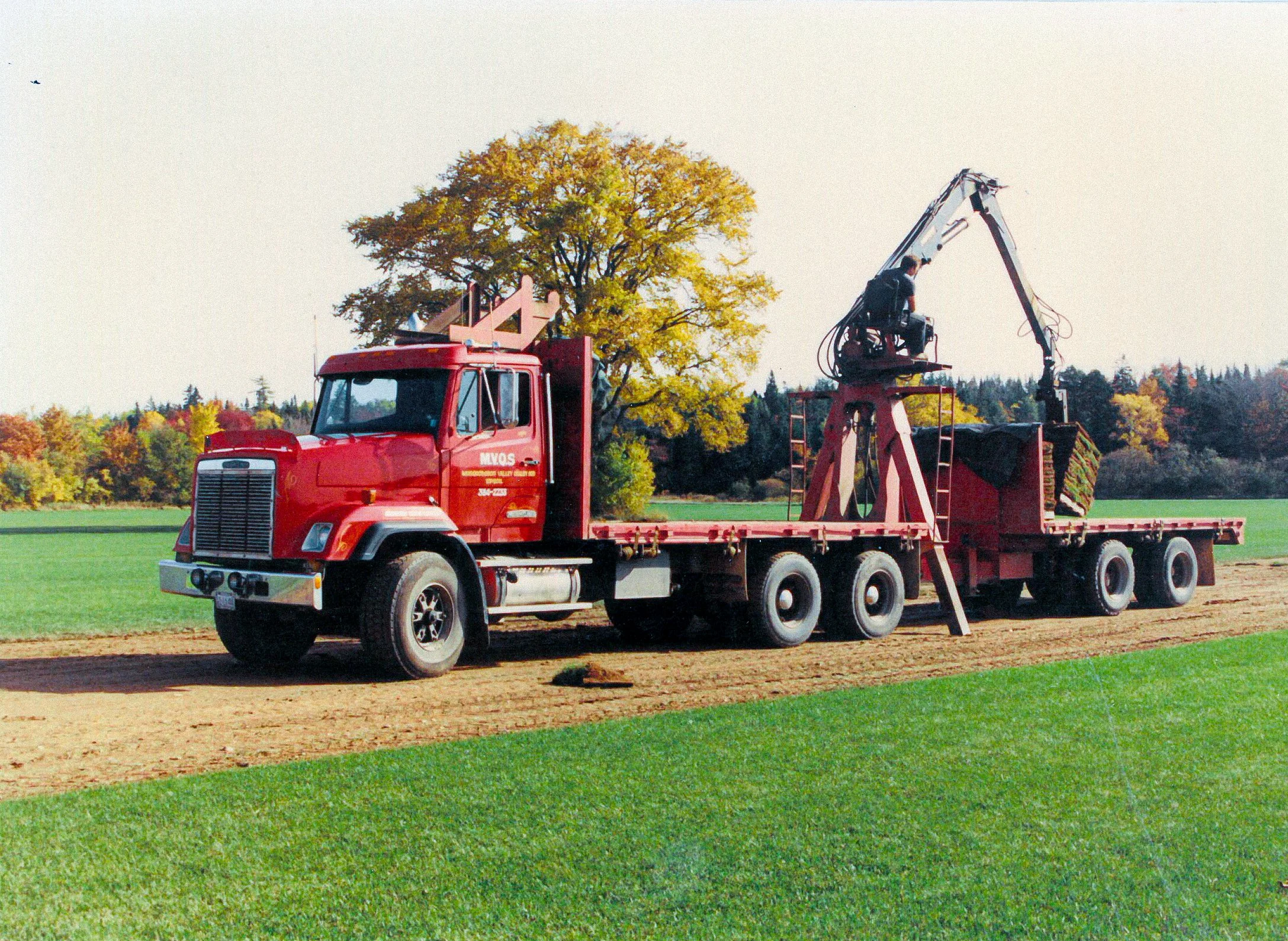 A red flatbed truck with a worker operating a crane attachment, set in a field with green grass and trees with autumn foliage in the background.