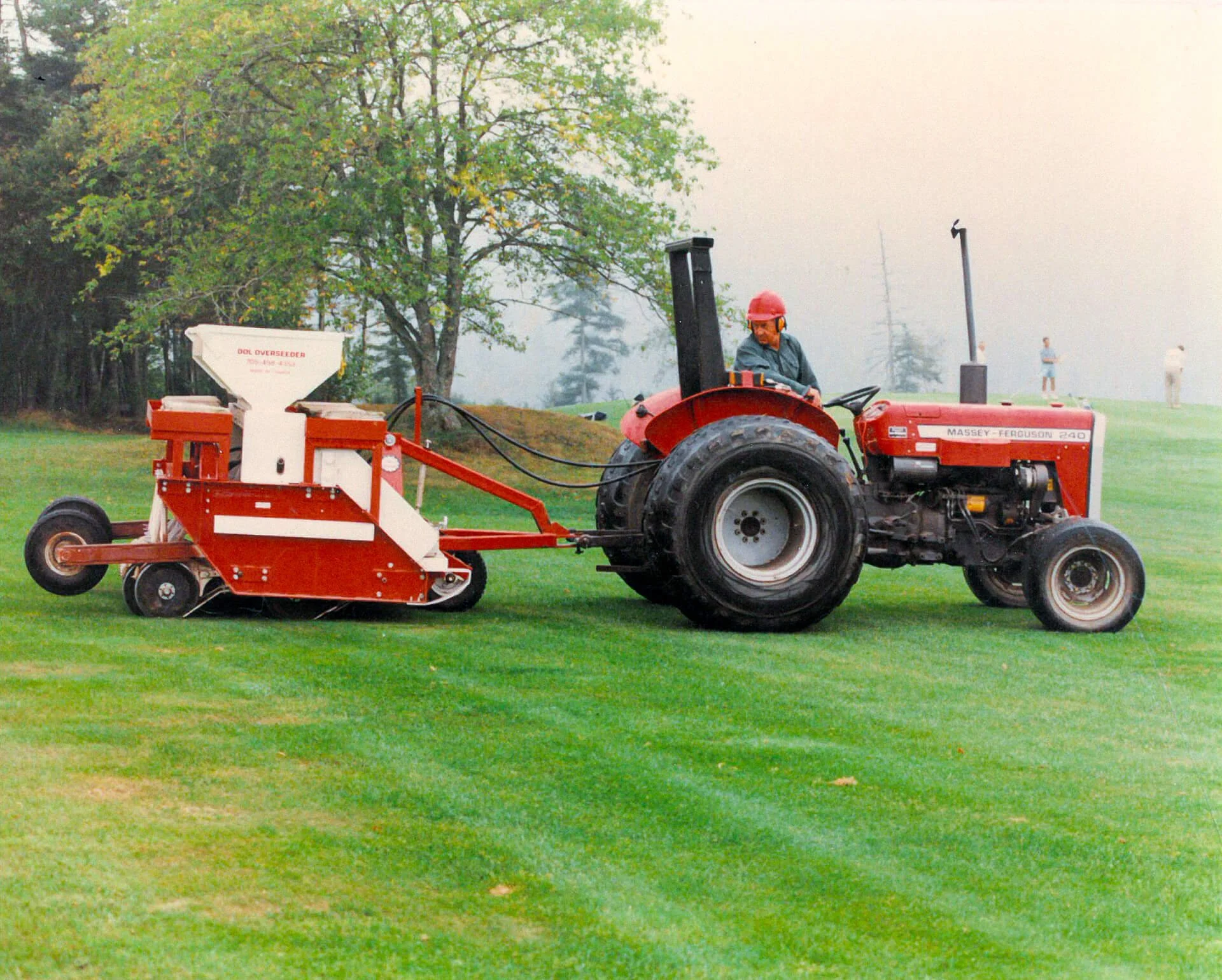 A man wearing a red helmet and glasses operating a red Massey Ferguson 240 tractor on a green lawn, with a tree and a few people in the background.