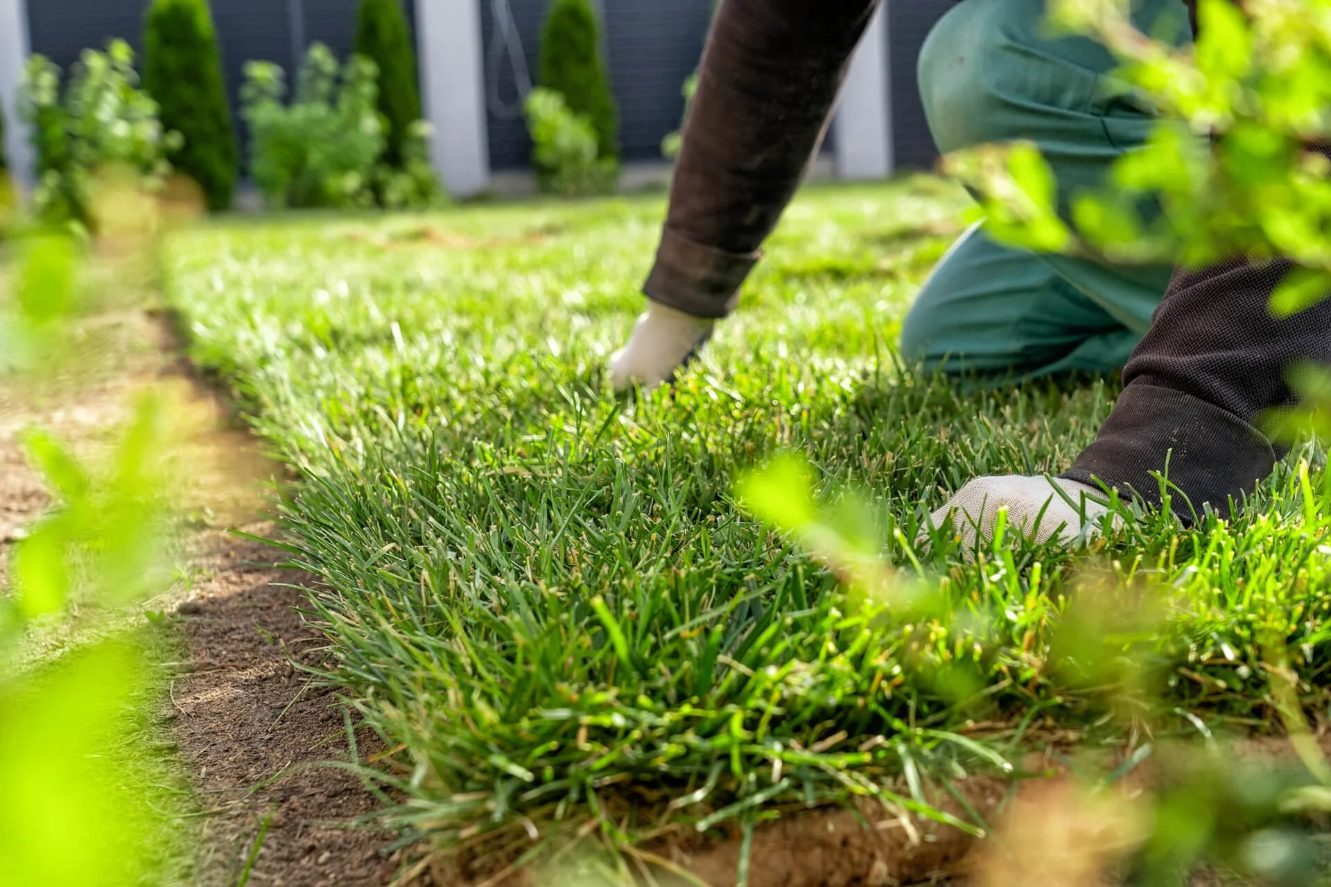 A person kneeling on the grass and planting or tending to plants in a garden. The individual is wearing gloves and teal clothing, with a focus on their hands and lower body.