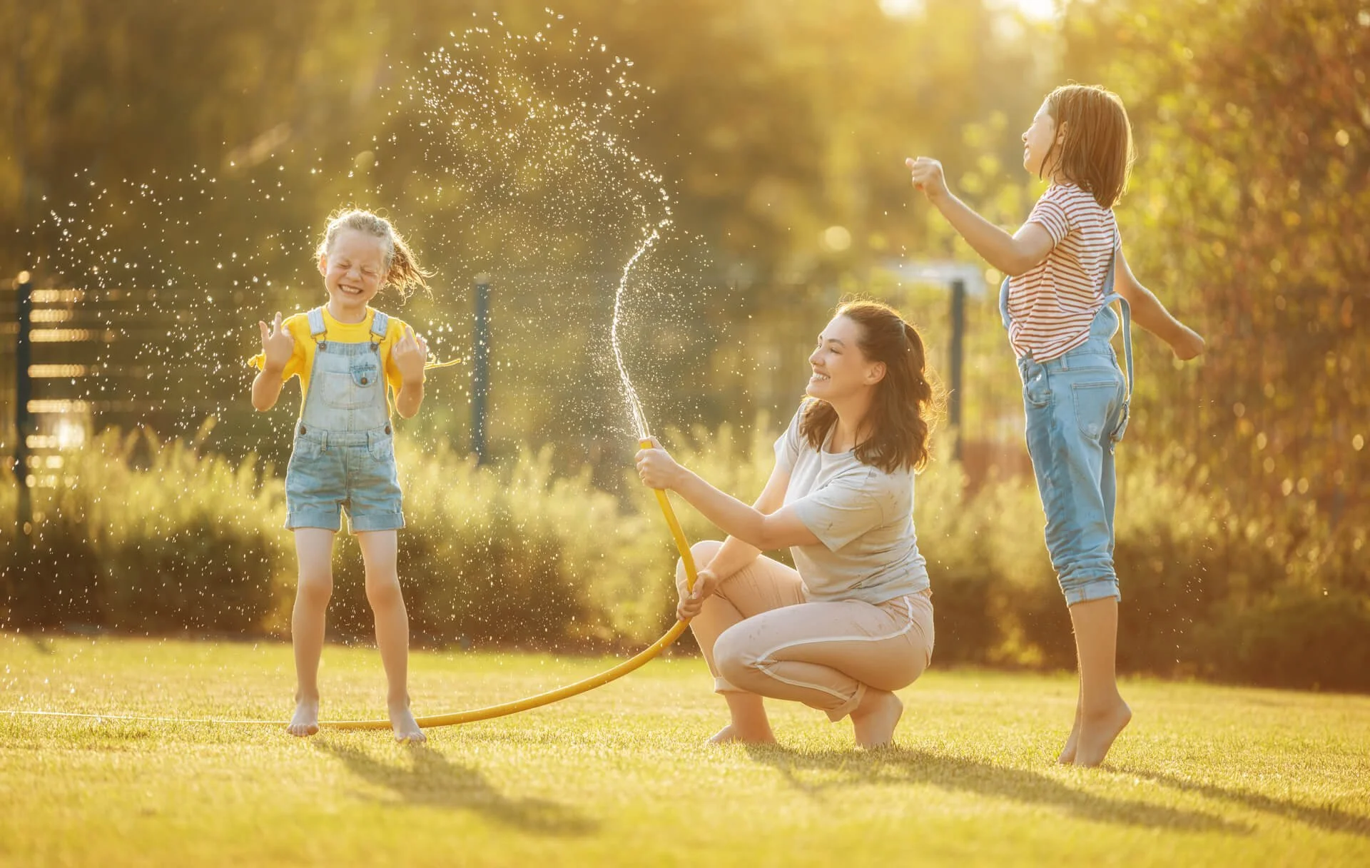 A woman and two young girls playing with a garden hose, spraying water, on a sunny day on their fresh sod with trees in the background.