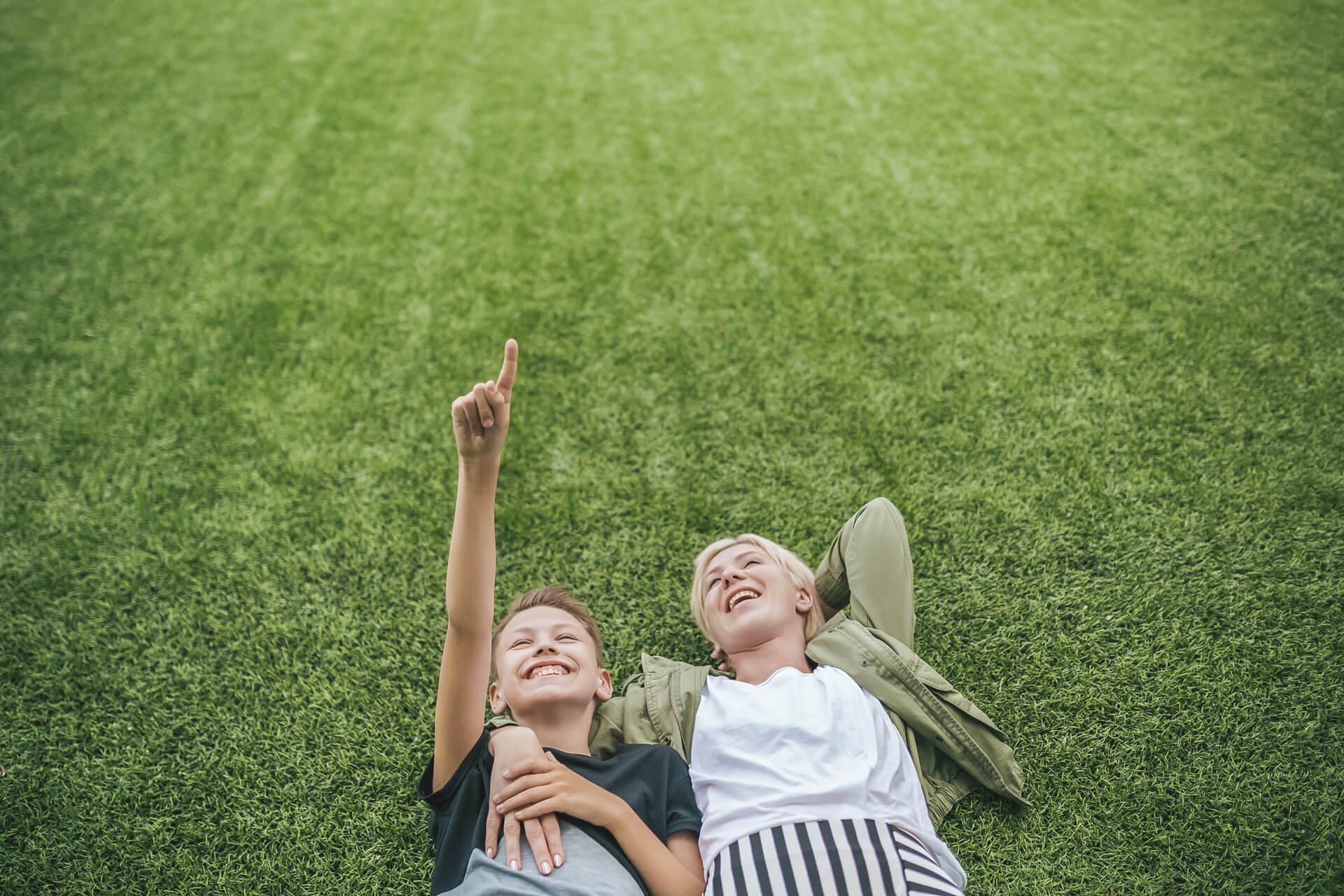 Two boys lying on green grass, smiling, with one boy pointing upward with his finger.