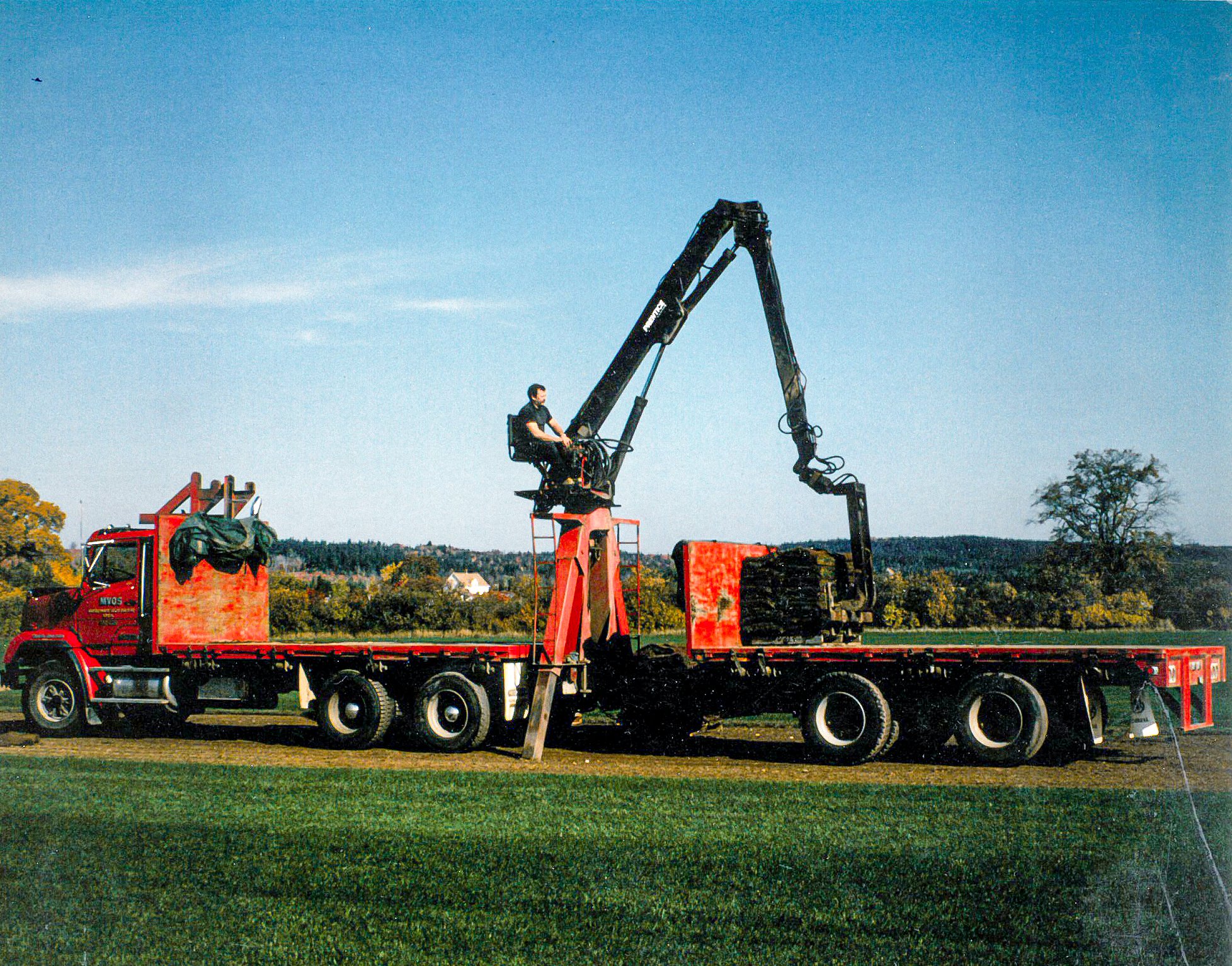 A person operating a crane mounted on a red truck in an open field, with trees and a few houses in the background under a clear sky.