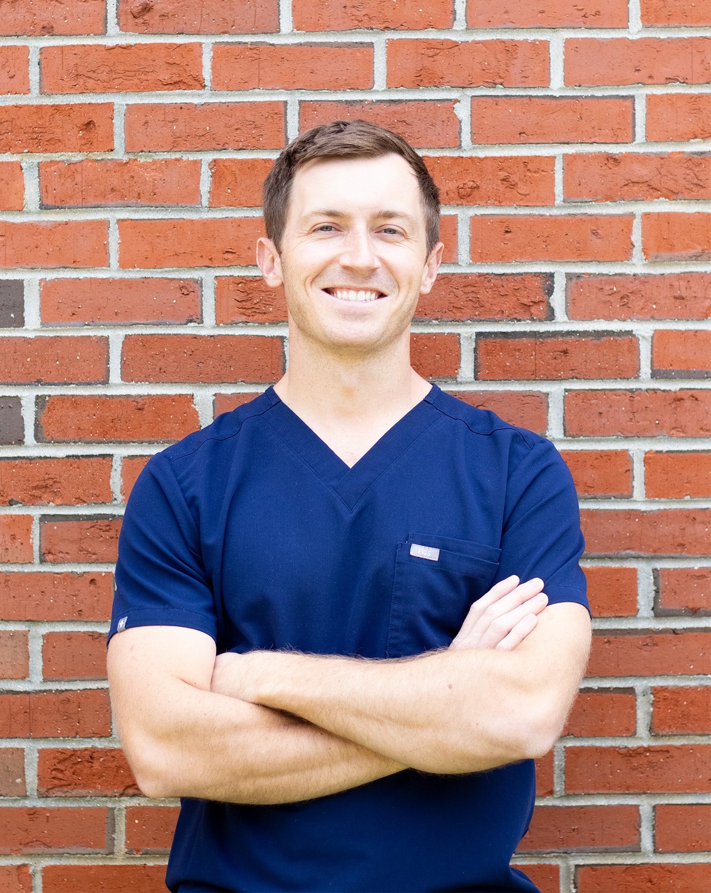 A smiling man with short brown hair and light skin, wearing a navy blue medical scrubs, standing cross-armed against a red brick wall.