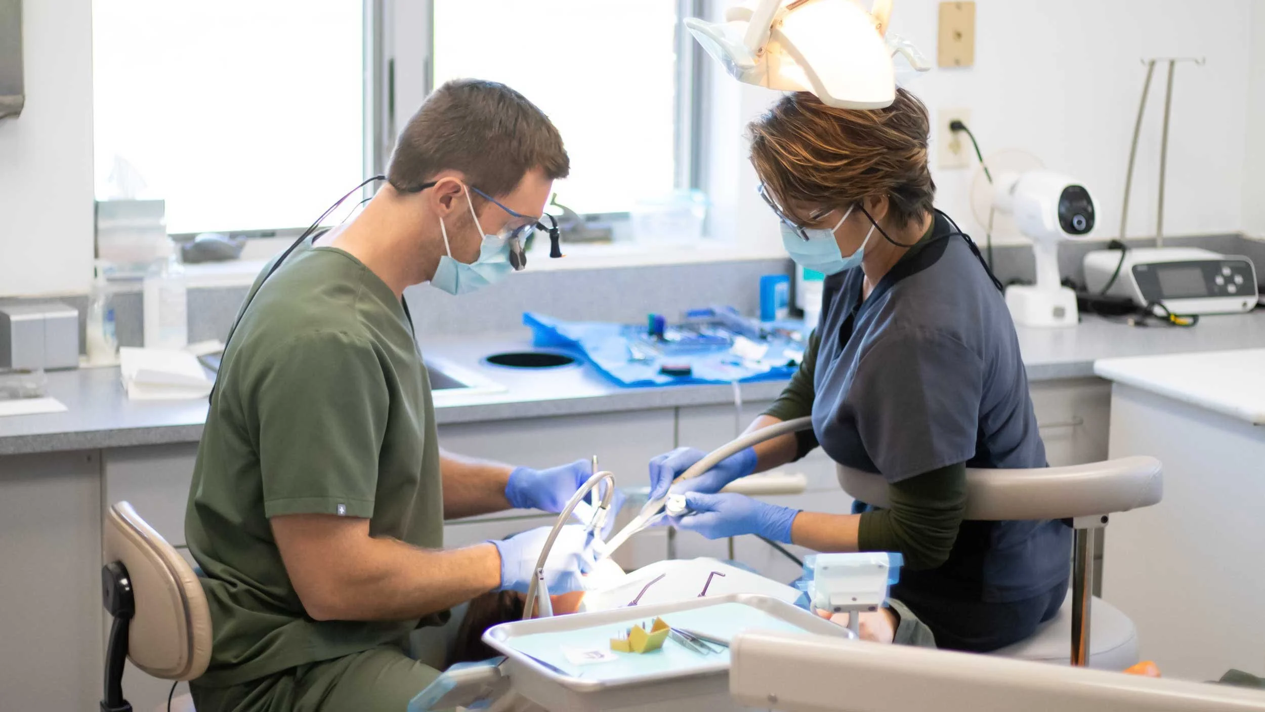 Two dental professionals, a man and a woman, wearing masks and gloves, performing a dental procedure in a clinical setting.