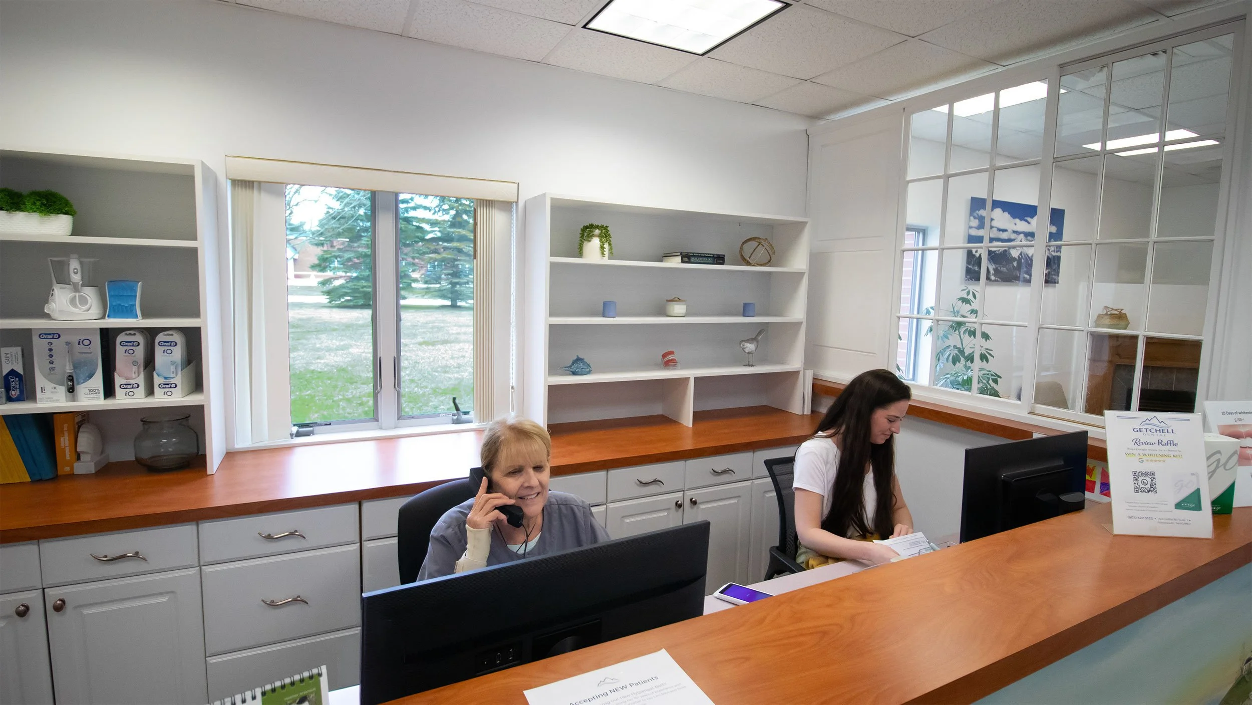 Office reception area with two women at a desk, one on the phone and the other reading, white shelves with books and decorations, large window showing outside trees.