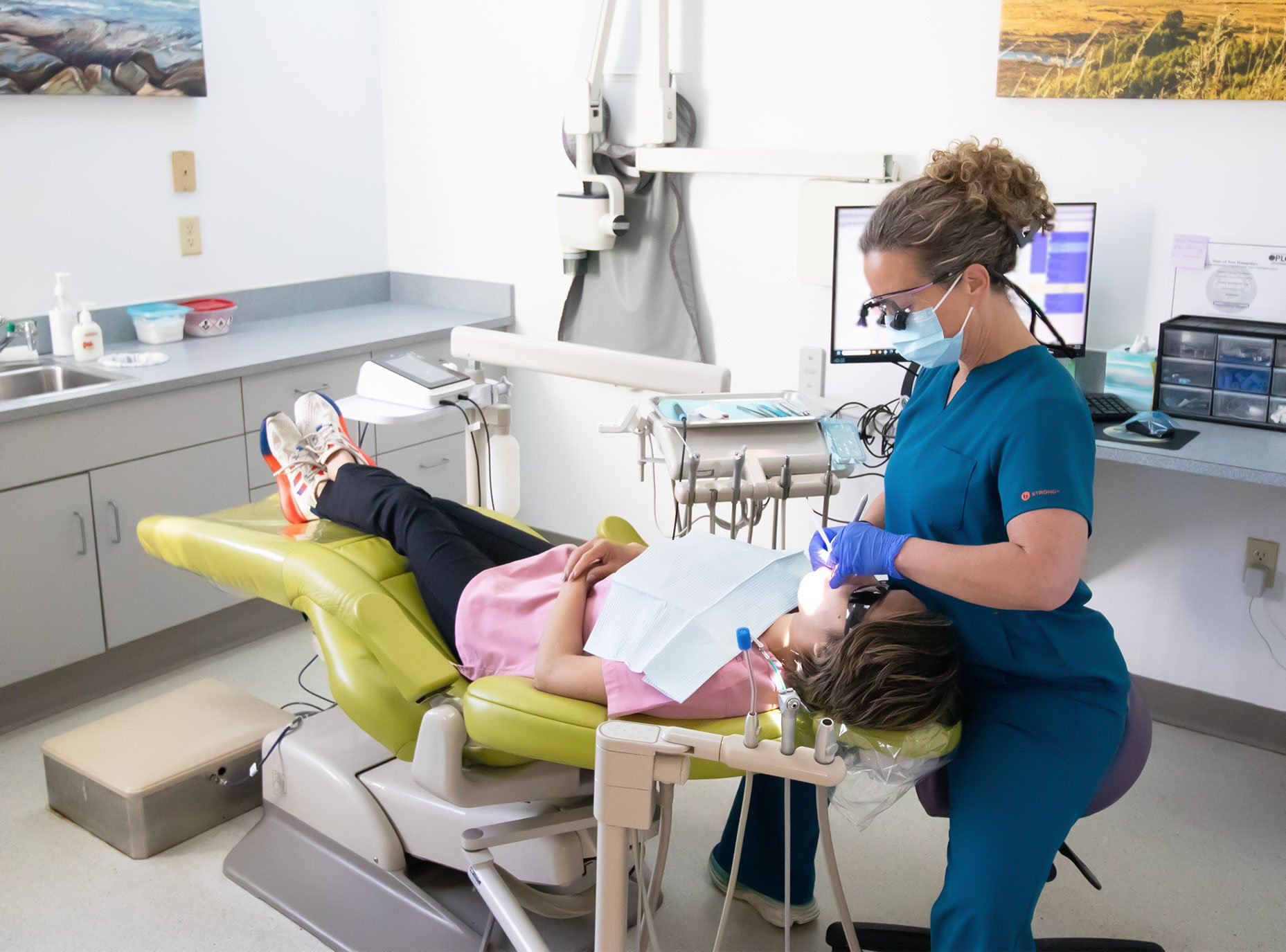 A dentist wearing blue scrubs, gloves, a mask, and magnifying glasses is working on a young patient lying in a dental chair. The patient has a pink shirt and is holding a book. Dental tools and equipment are visible in the clinic, with a monitor displaying dental images in the background.