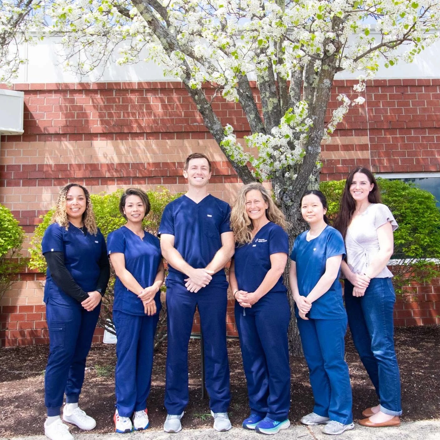 A group of six healthcare professionals standing outside in front of a flowering tree and brick wall, wearing blue scrubs and casual clothing, smiling at the camera.
