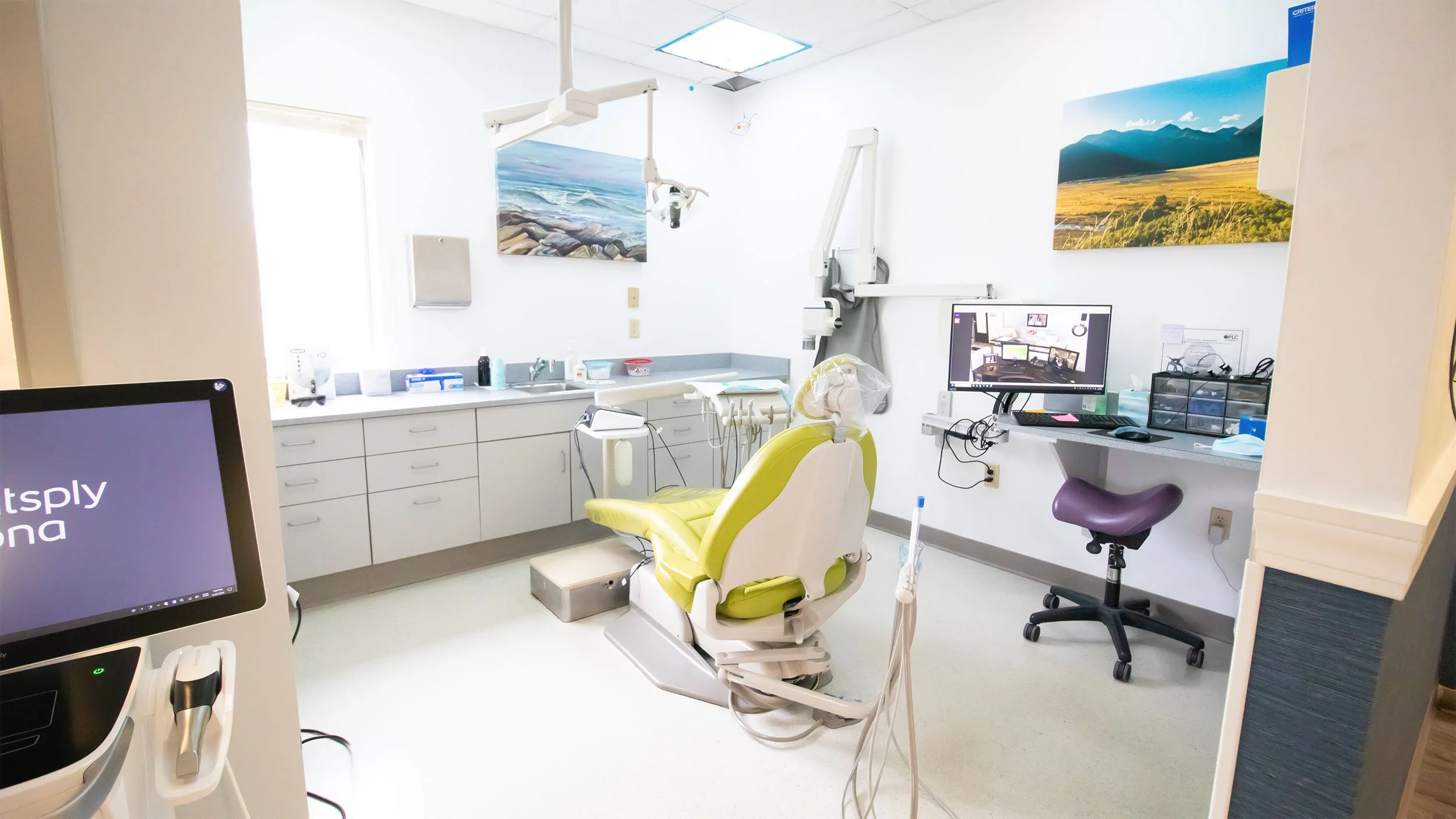 A dentist's office with dental chair, computer monitors, paintings of landscapes, dental tools, and medical supplies, brightly lit and organized.