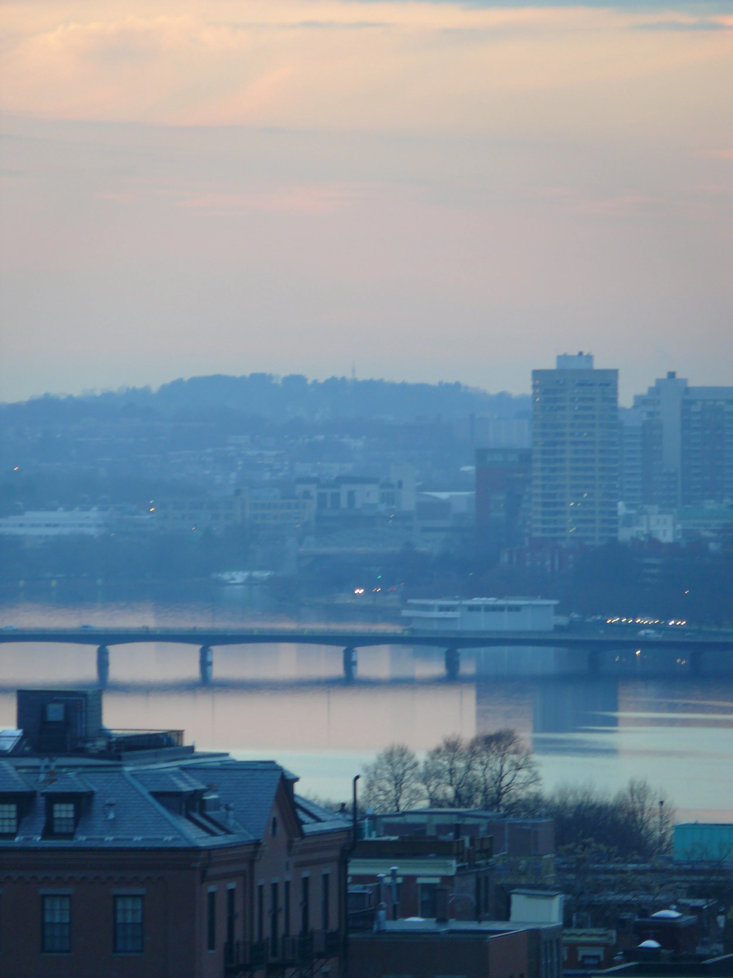 Boston/Cambridge view of the Charles River with rooftops, a bridge, water, distant buildings, hills, and a cloudy sky at dusk or dawn.