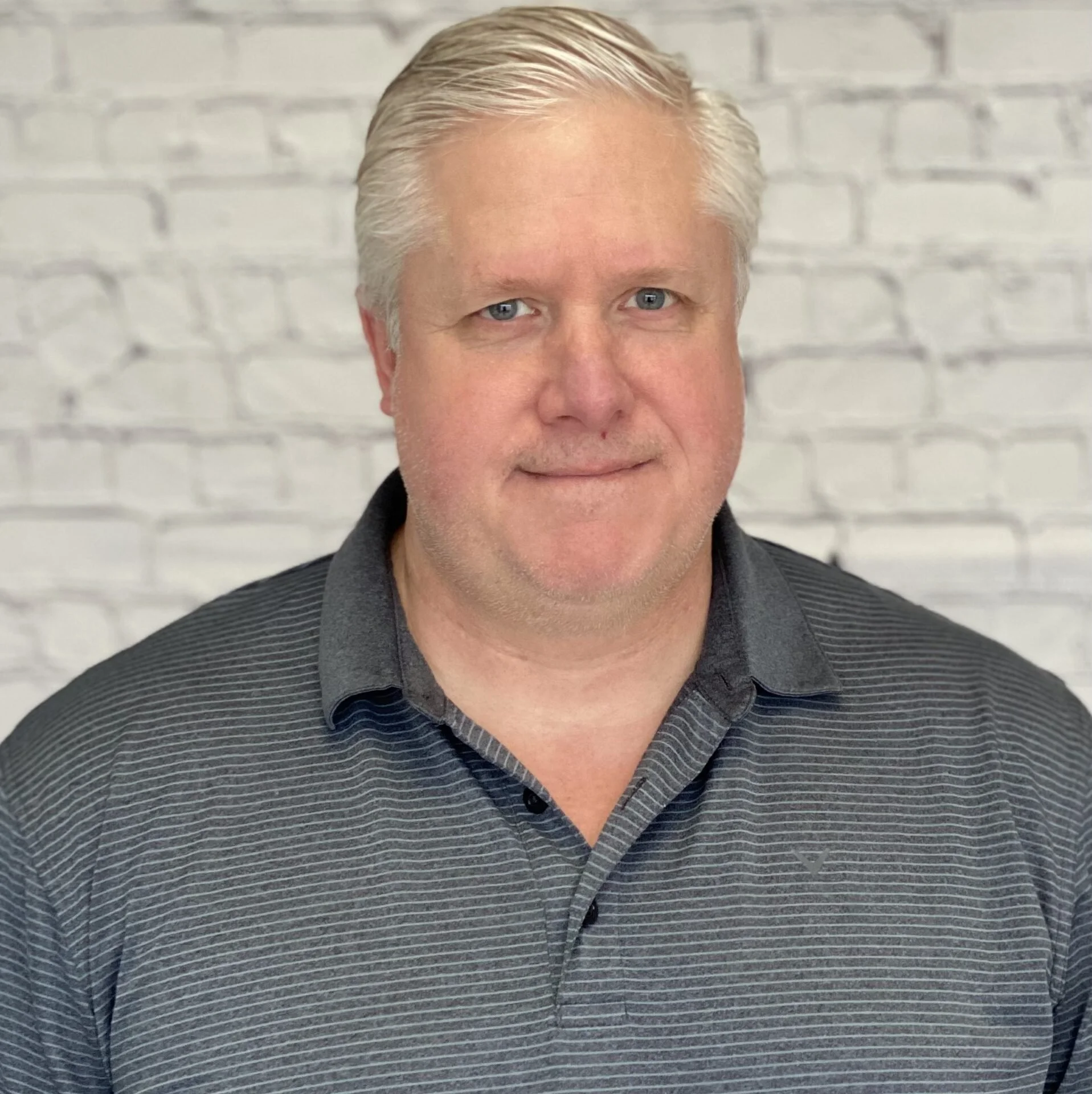 A middle-aged man with light skin, blonde hair styled back, and blue eyes, wearing a dark collared shirt with thin white stripes, standing against a white brick wall.