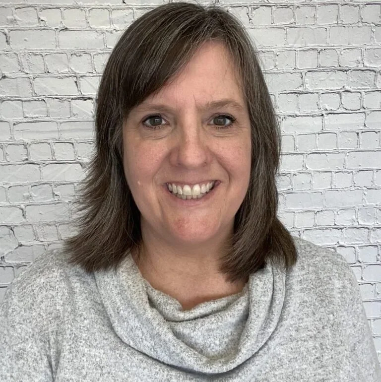 A woman with shoulder-length brown hair smiling, wearing a gray cowl neck sweater, standing in front of a white brick wall.