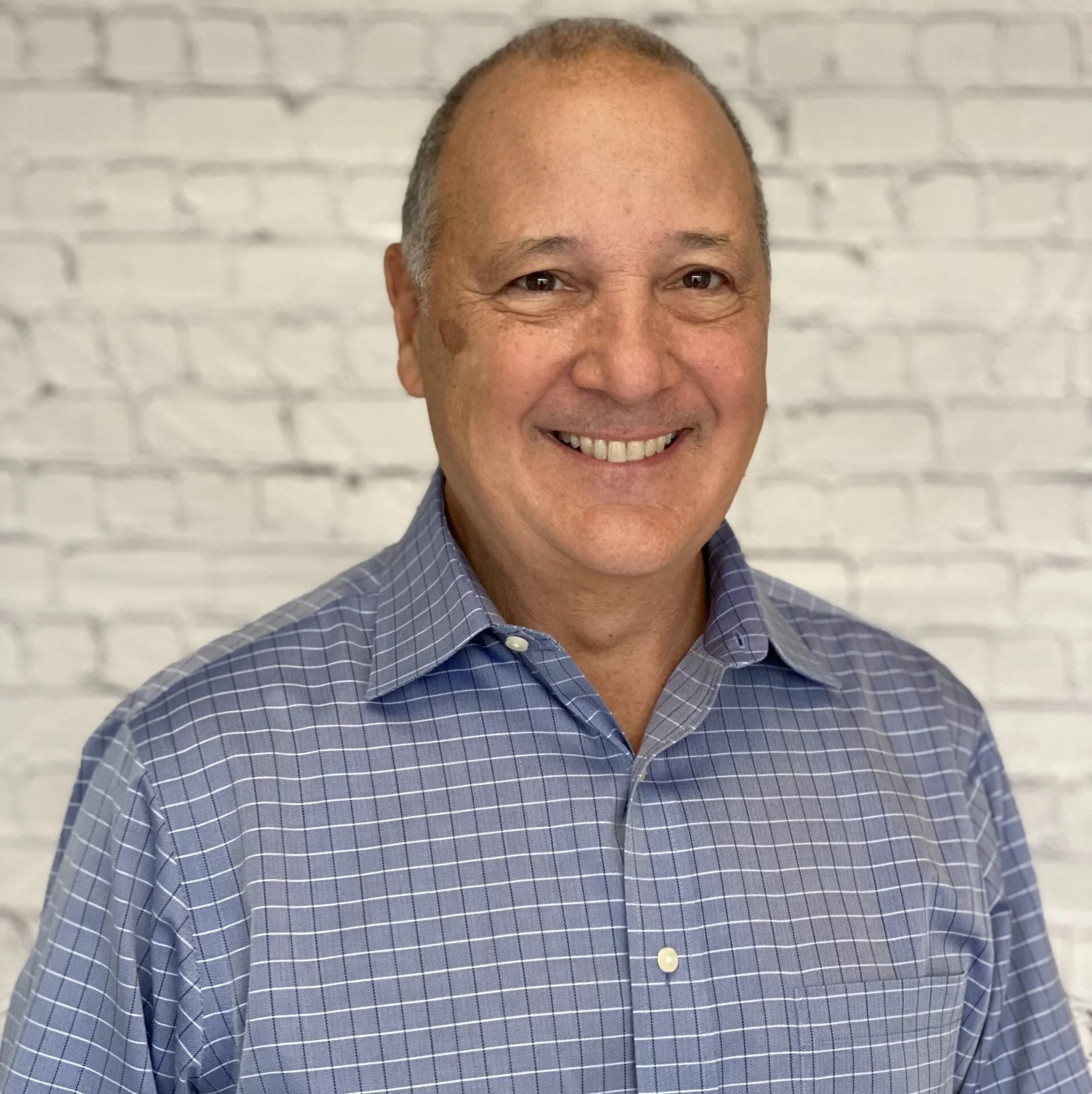 A smiling man with a shaved head wearing a blue checkered dress shirt, standing in front of a white brick wall.