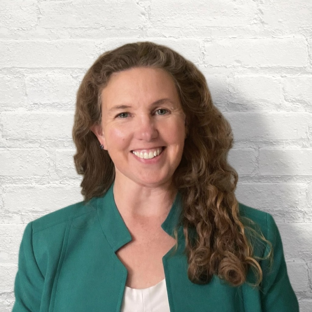 A woman with long, curly brown hair smiling in front of a white brick wall, wearing a teal blazer over a white top.