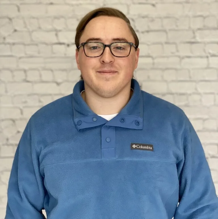 A young man with glasses, light skin, and brown hair standing in front of a white brick wall, wearing a blue Columbia fleece jacket.