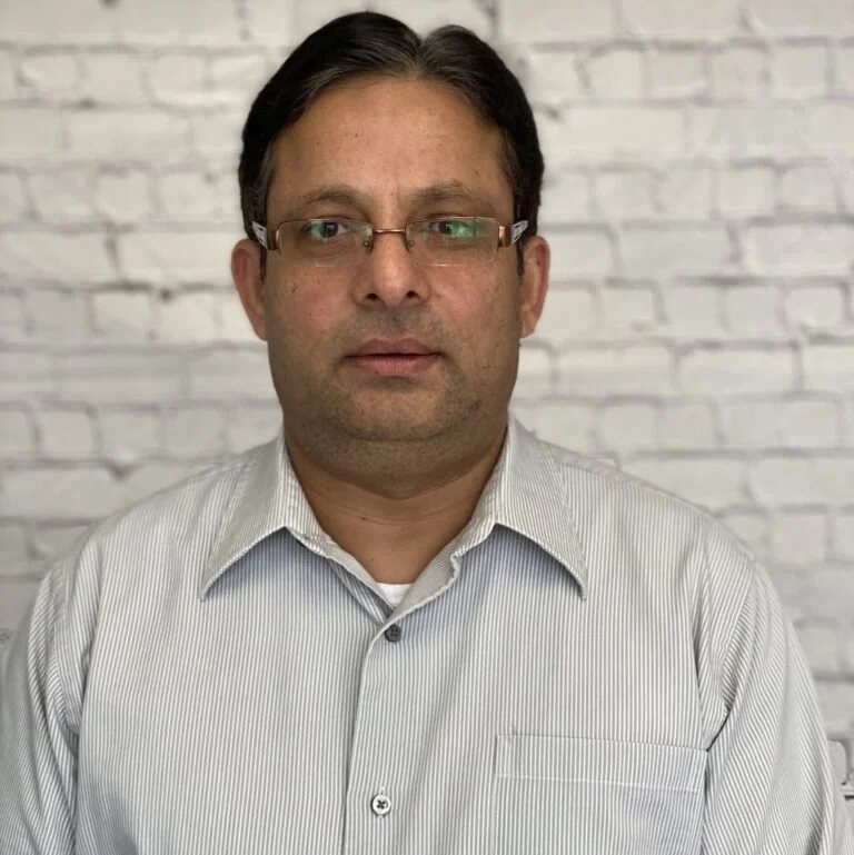 A man with dark hair, glasses, and a serious expression wearing a light gray, striped button-up shirt standing in front of a white brick wall