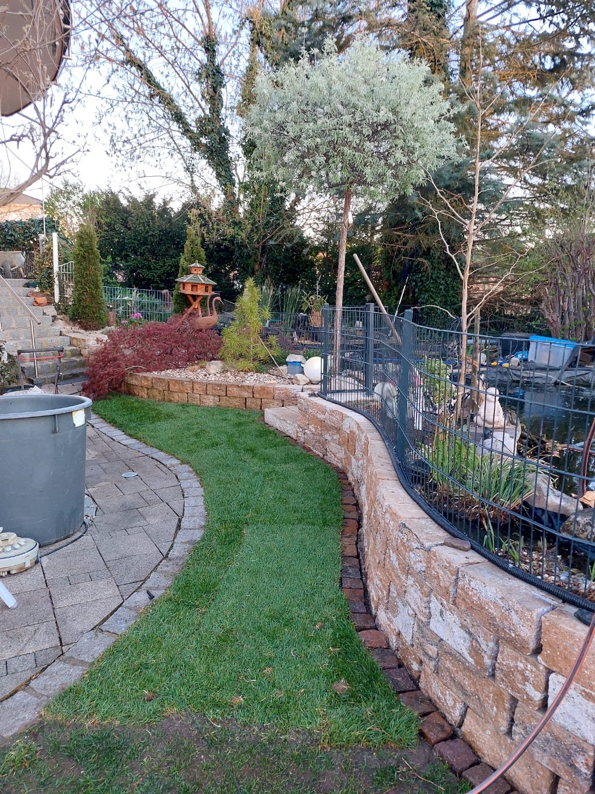 Garten mit Steinmauer, Bäumen, Sträuchern, einer kleinen Statue, Gartenpavillon und einem Wasserbecken.