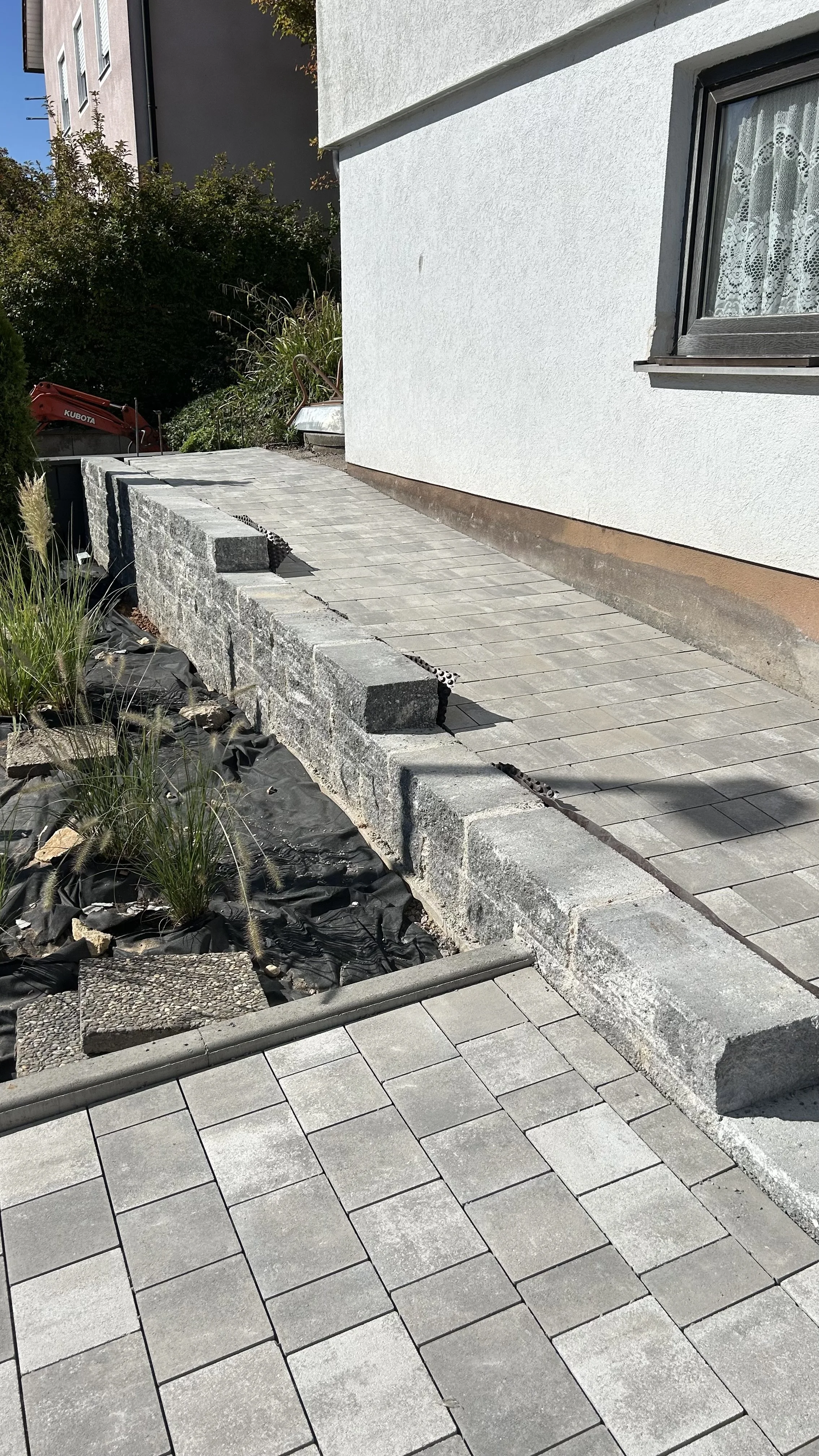 Das Bild zeigt einen Außenbereich vor einem Haus mit einer neu verlegten Gehwegplatte aus grauen Steinen. Im Vordergrund sind frisch verlegte Pflastersteine, im Hintergrund sieht man den angestrebten Rand und eine Mauer aus Steinen. Es gibt eine weiße Hauswand mit einem Fenster, das durch eine Gardine verdeckt ist. Auf der linken Seite sind Pflanzen und eine kleine Baustelle mit Baumaterialien und einer Gartenbrücke sichtbar.