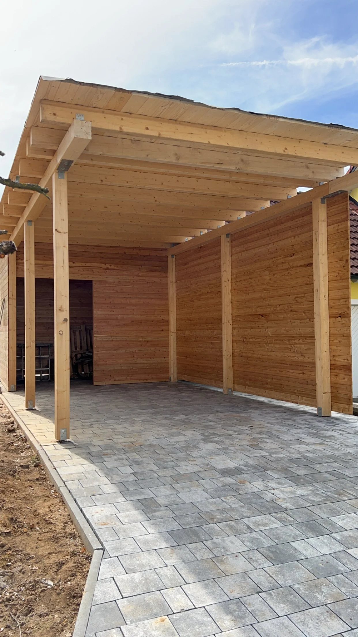 Ein Carport oder Unterstand wird gebaut, mit Holzrahmen und Holzverkleidung. Der Boden ist mit Pflastersteinen gepflastert, und der Himmel ist bläulich mit einigen Wolken.