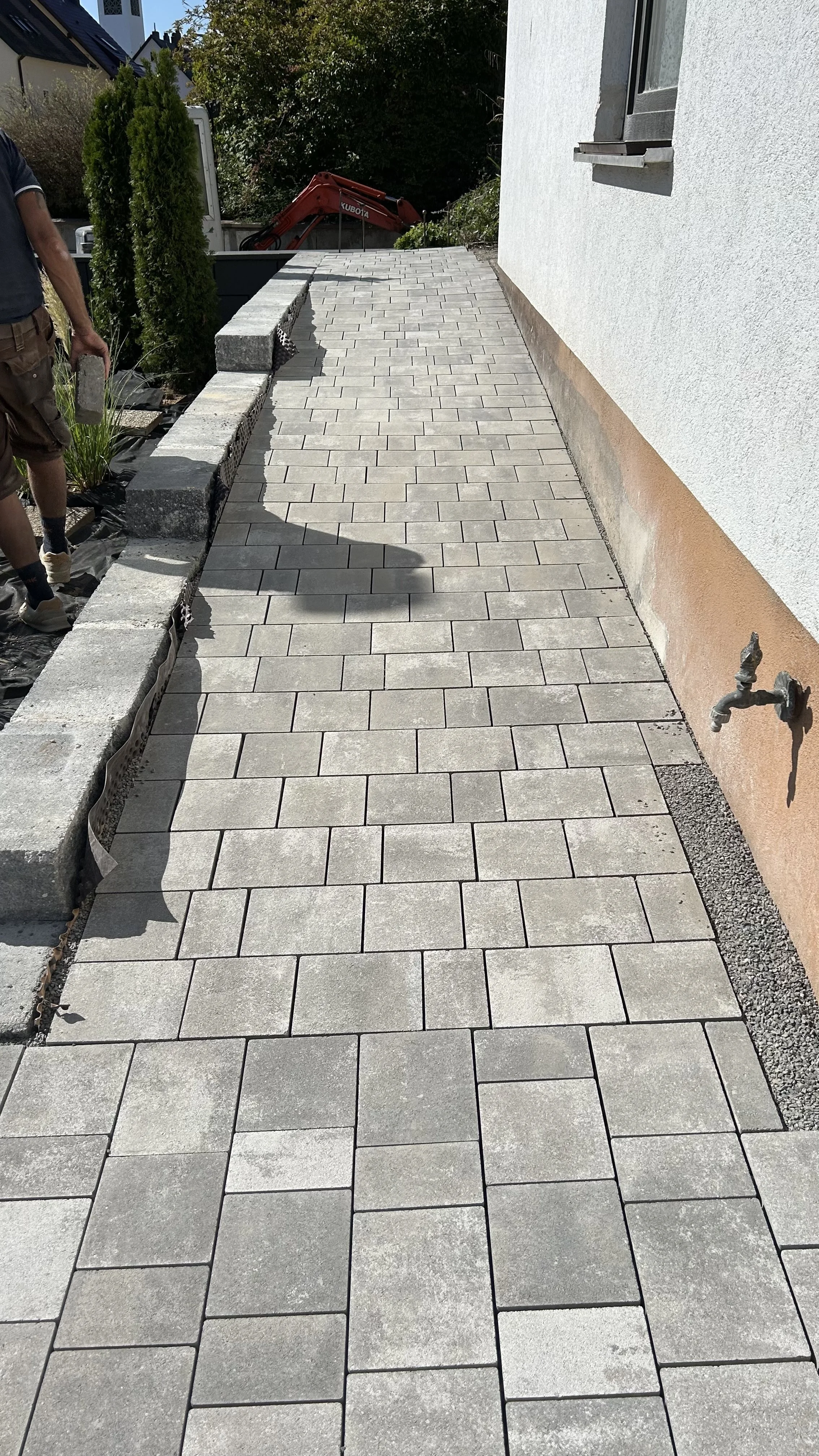 Der Bild zeigt einen Gehweg, auf dem Pflastersteine verlegt werden. Auf der linken Seite sind Bauarbeiten im Gange, und ein Mann trägt Baumaterial. Im Hintergrund sind Bäume und ein Kran sichtbar.