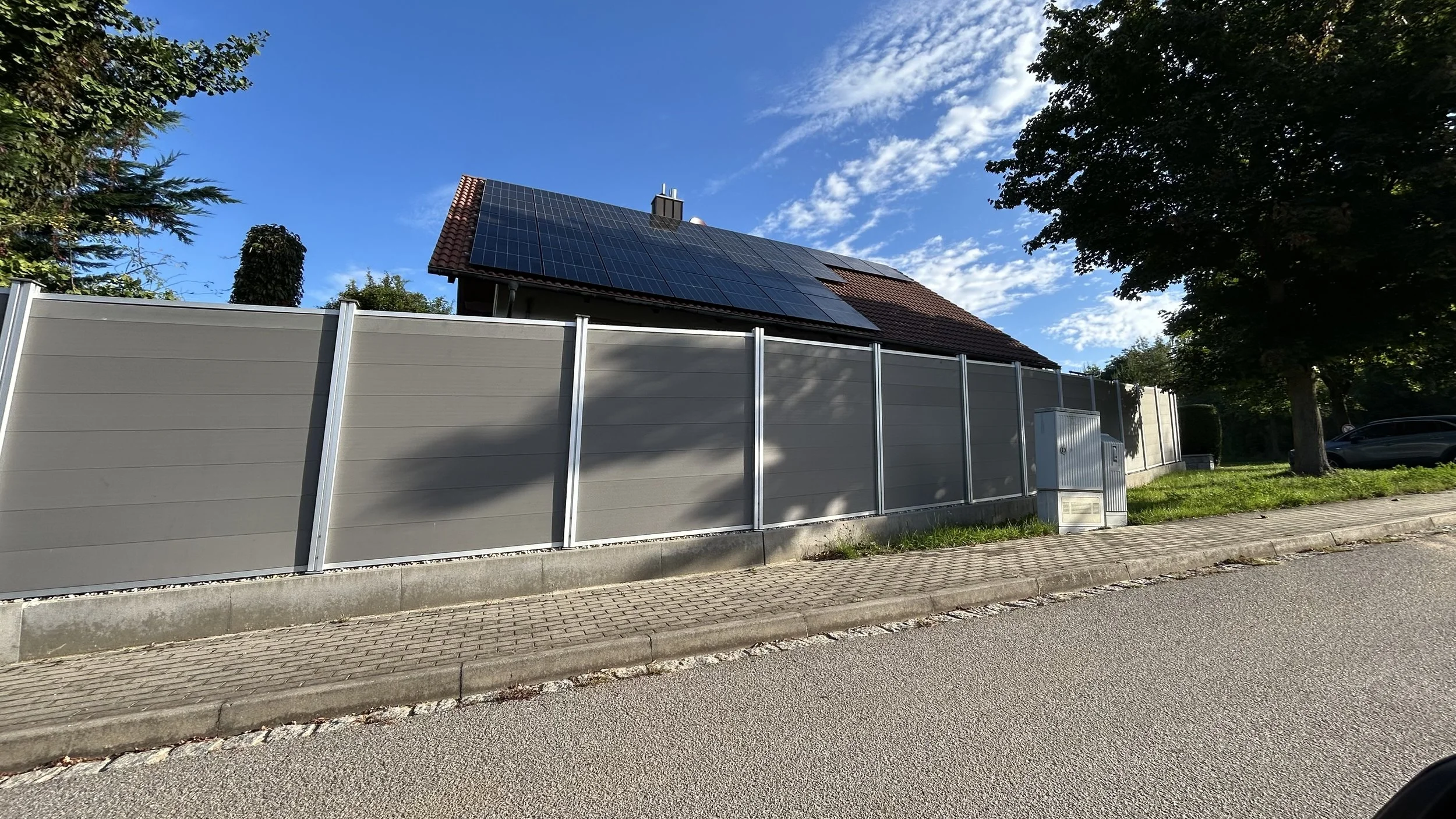 Haus mit Dachmodernisierung durch Solarpanele auf dem Dach, umgeben von Bäumen und einer Mauer, bei sonnigem Himmel.