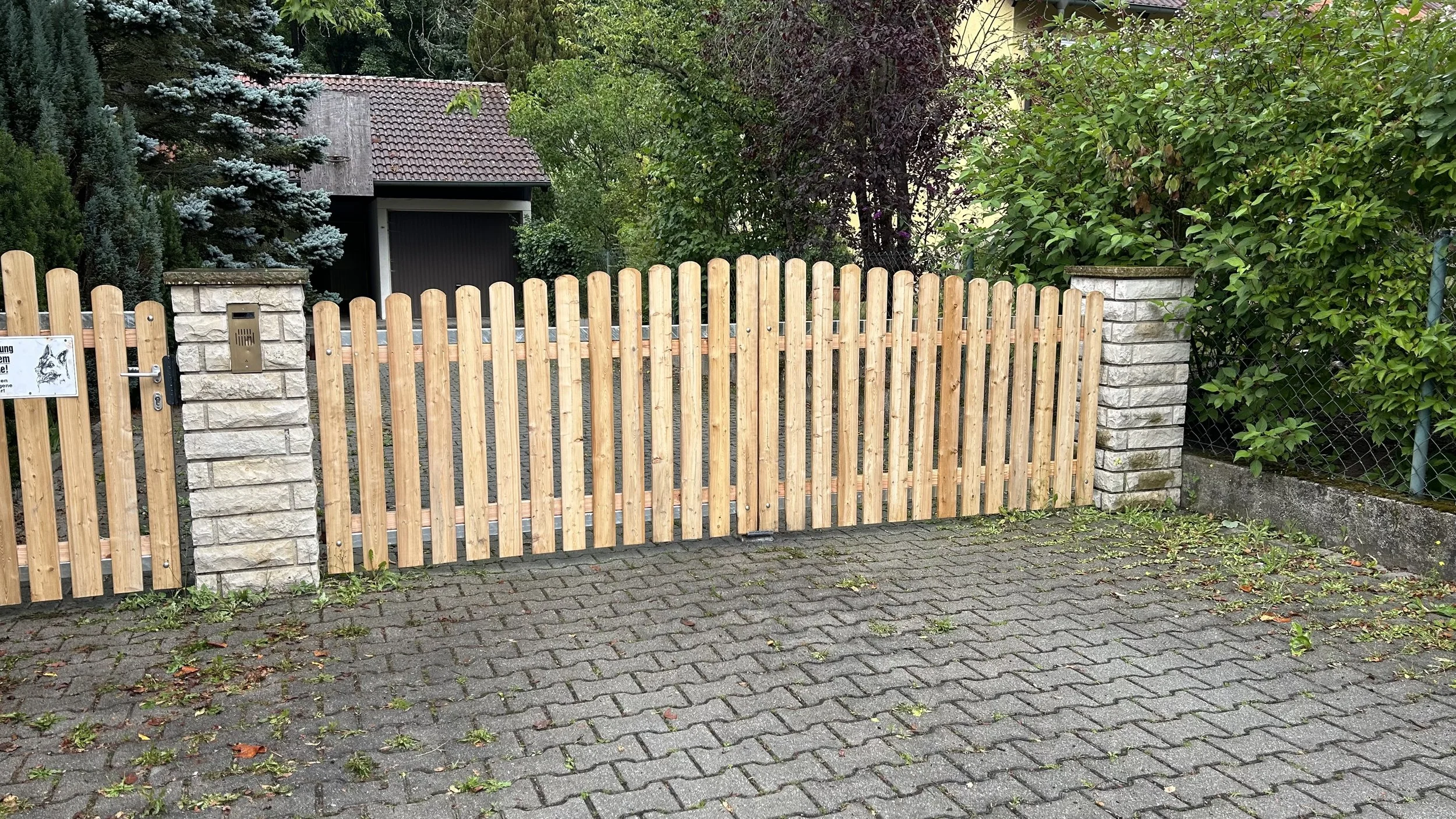 Holzzaun vor einer Steinmauer mit Türschloss, umgeben von grünen Büschen und Bäumen, im Vordergrund eine gepflasterte Straße.