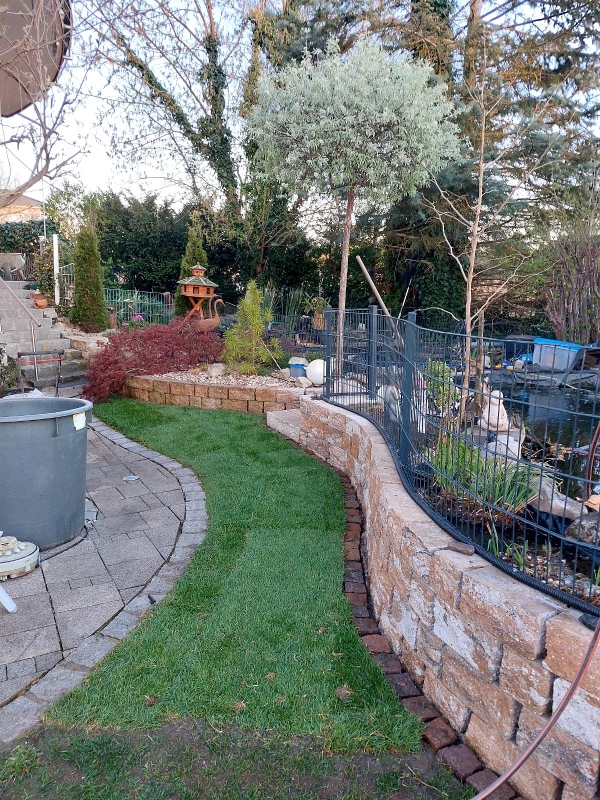 Ein Garten mit Steinstufen, einem kleinen Wasserbecken, einem Zaun, Bäumen, Sträuchern und Gartendekorationen
