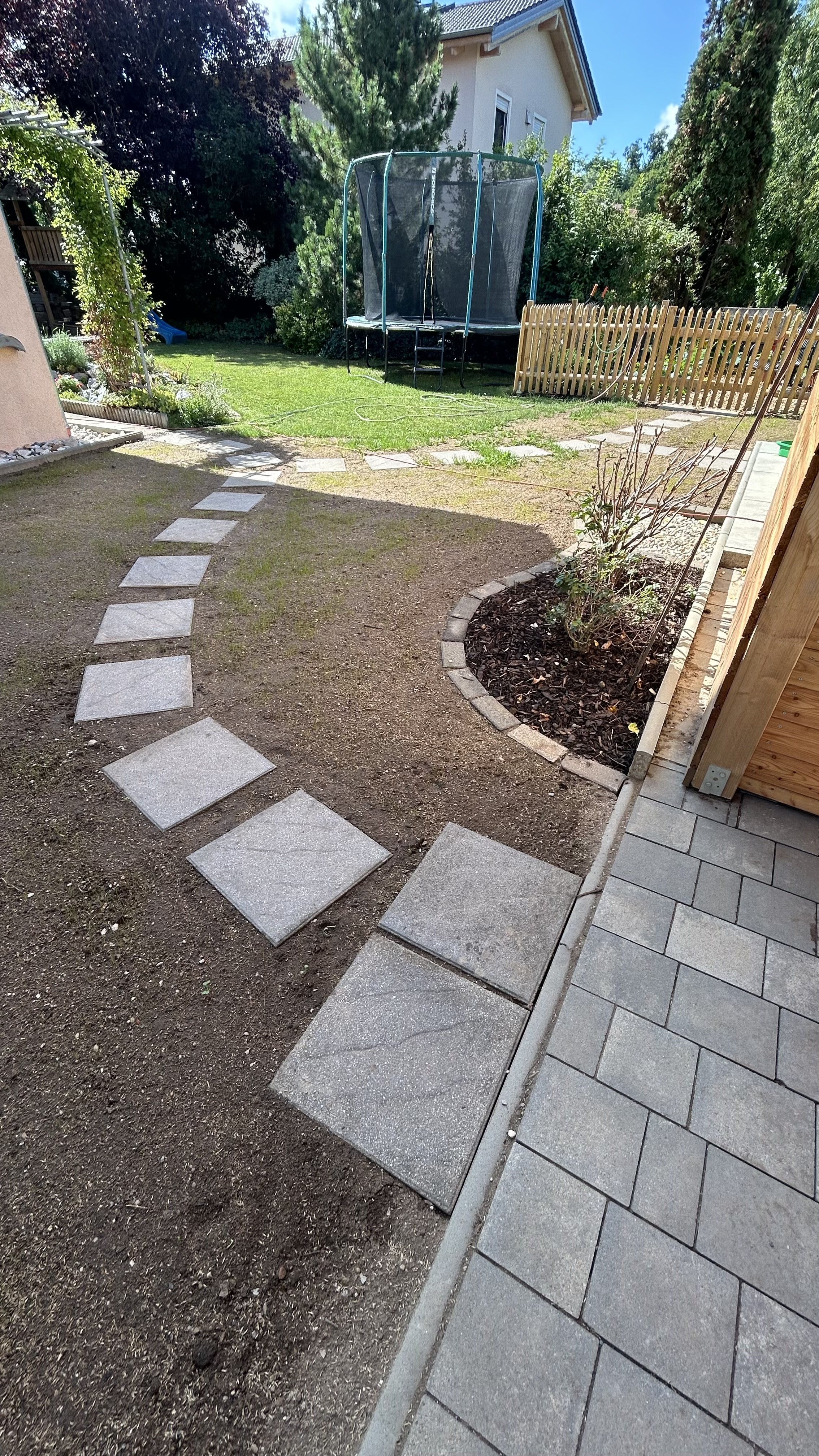 Der Garten zeigt einen gepflasterten Weg mit Quadraten, einen Rasenbereich mit einem Trampolin, Blumenbeete und einen Zaun an der Seite.