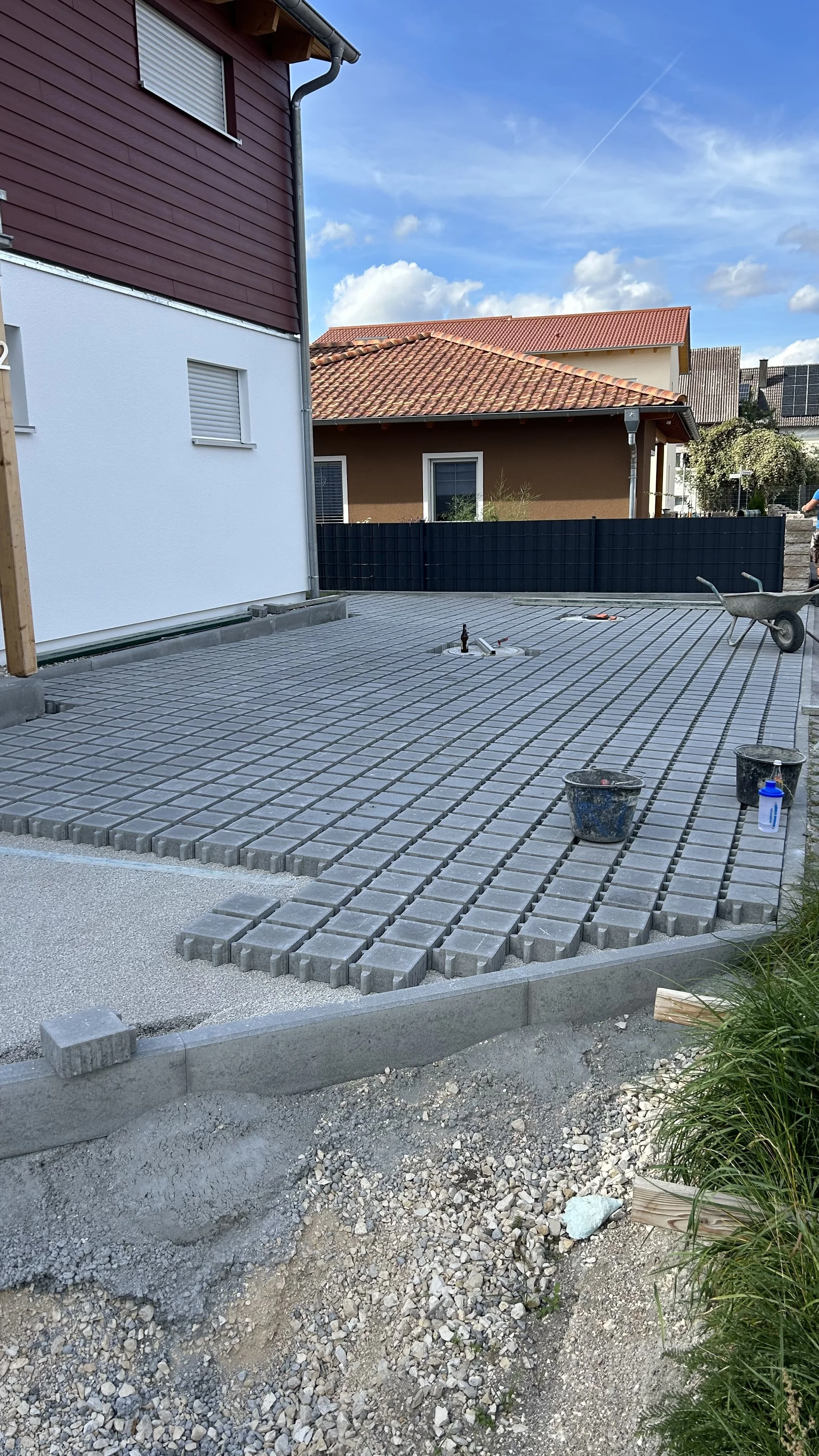 Der Eingangsbereich eines Hauses wird mit Pflastersteinen ausgelegt, mit Werkzeugen und Baumaterialien für die Pflasterung, neben einem Wohnhaus mit rotem Dach und weißer Wand.