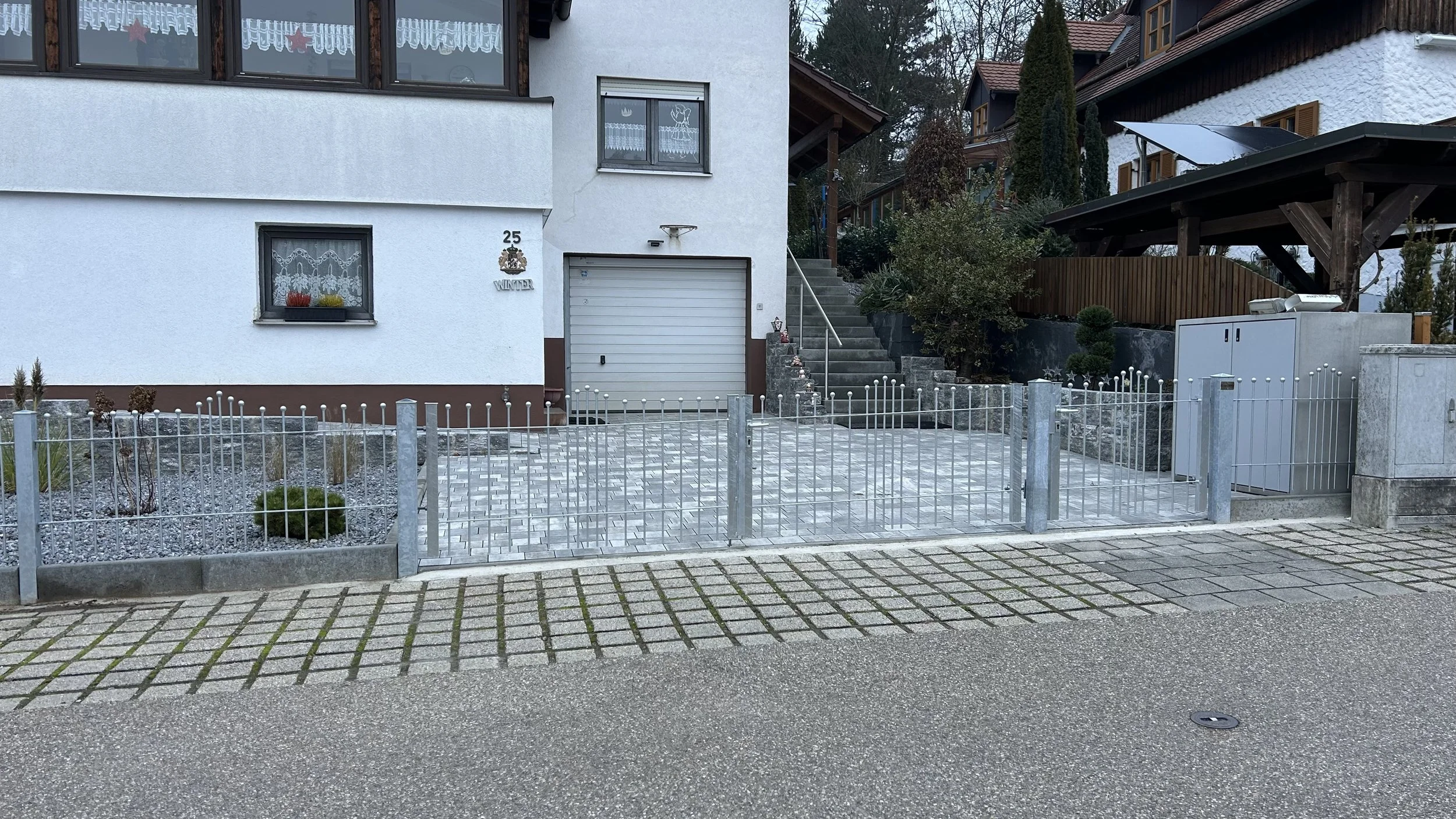 Ein Haus mit weißer Fassade, einer Garage, Treppen und Gärtnerarbeiten im Vorgarten, umgeben von einem Metallzaun.