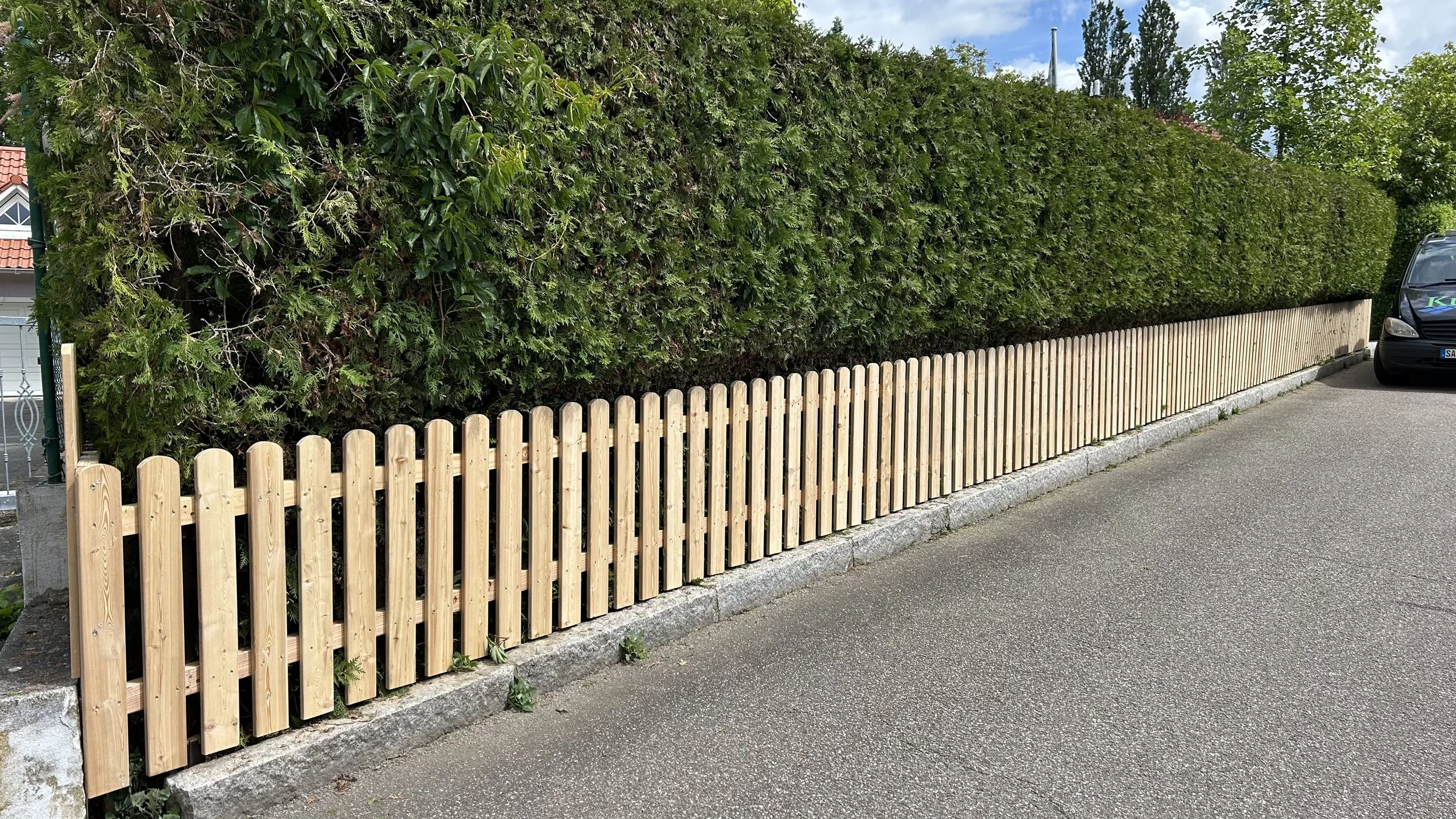 Ein Holzzaun, der eine dichte grüne Hecke entlang eines Gehwegs begrenzt. Auf der rechten Seite ist ein schwarzes Fahrzeug zu sehen.