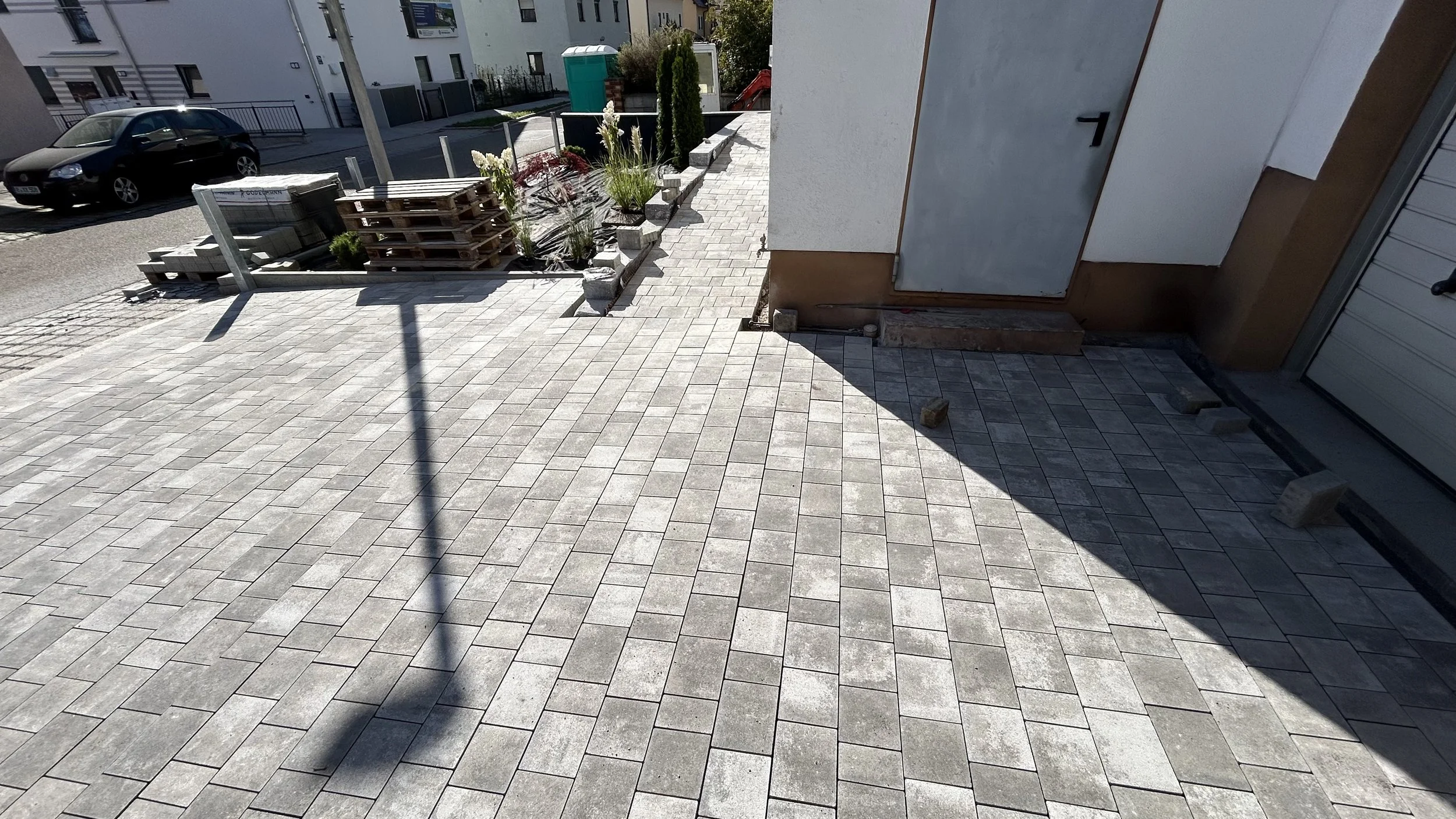 Ein Gehweg mit Pflastersteinen vor einem Gebäude, im Hintergrund geparkte Autos, und ein Gartenbereich mit Pflanzen und Paletten. Es ist sonnig und es gibt einen Schatten im Vordergrund.